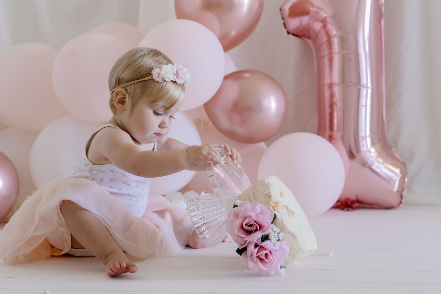Baby in white dress playing with roses amidst pink party decorations and balloons.