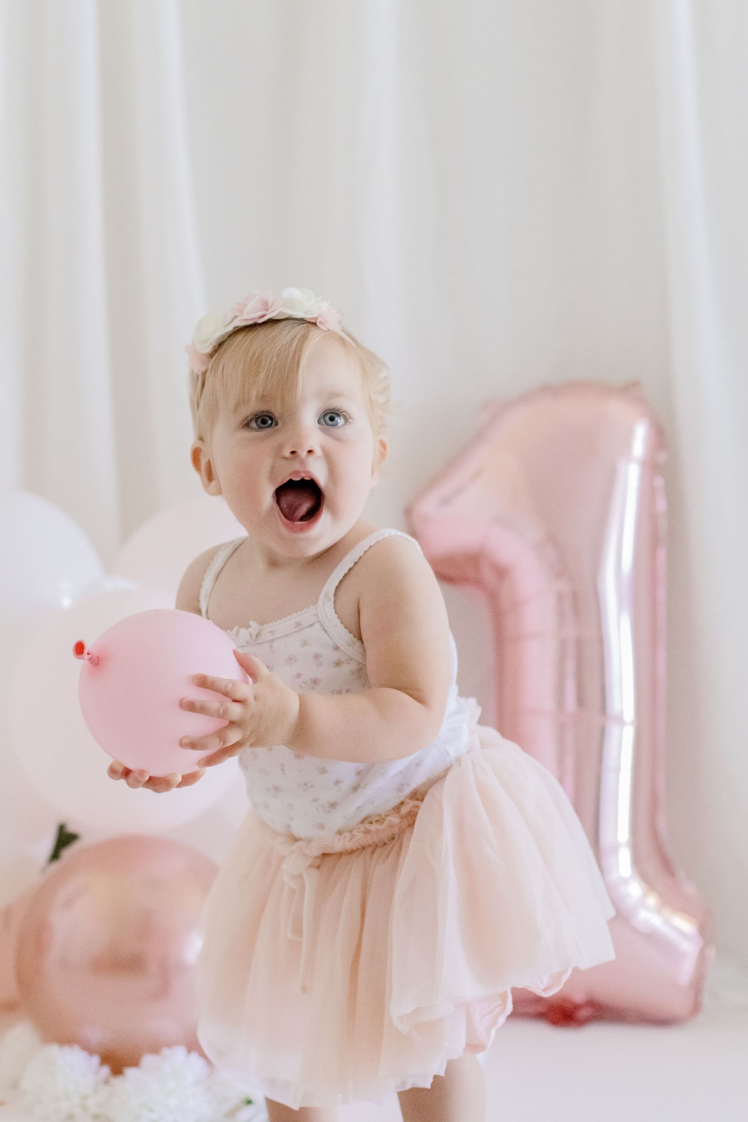 Toddler in tulle dress expressing joy while holding pink balloons at a first birthday photoshoot.