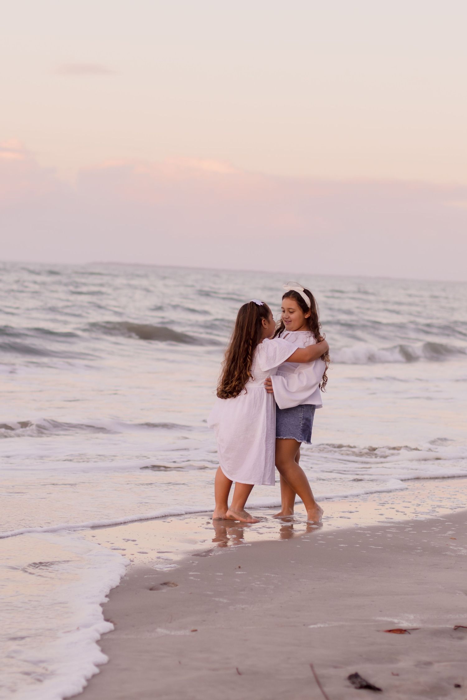 Two people embrace while standing ankle-deep in ocean waves during a peaceful sunset on the beach.