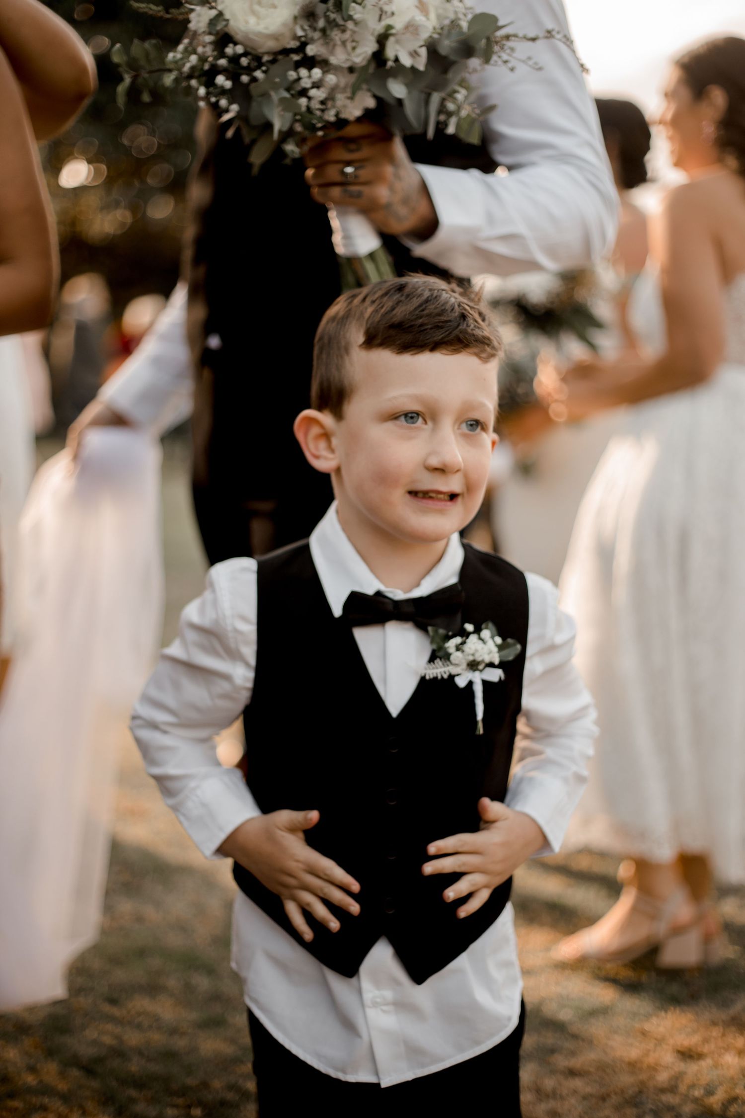 A young boy in formal black vest and bowtie at a wedding ceremony, with white flowers pinned to his lapel.