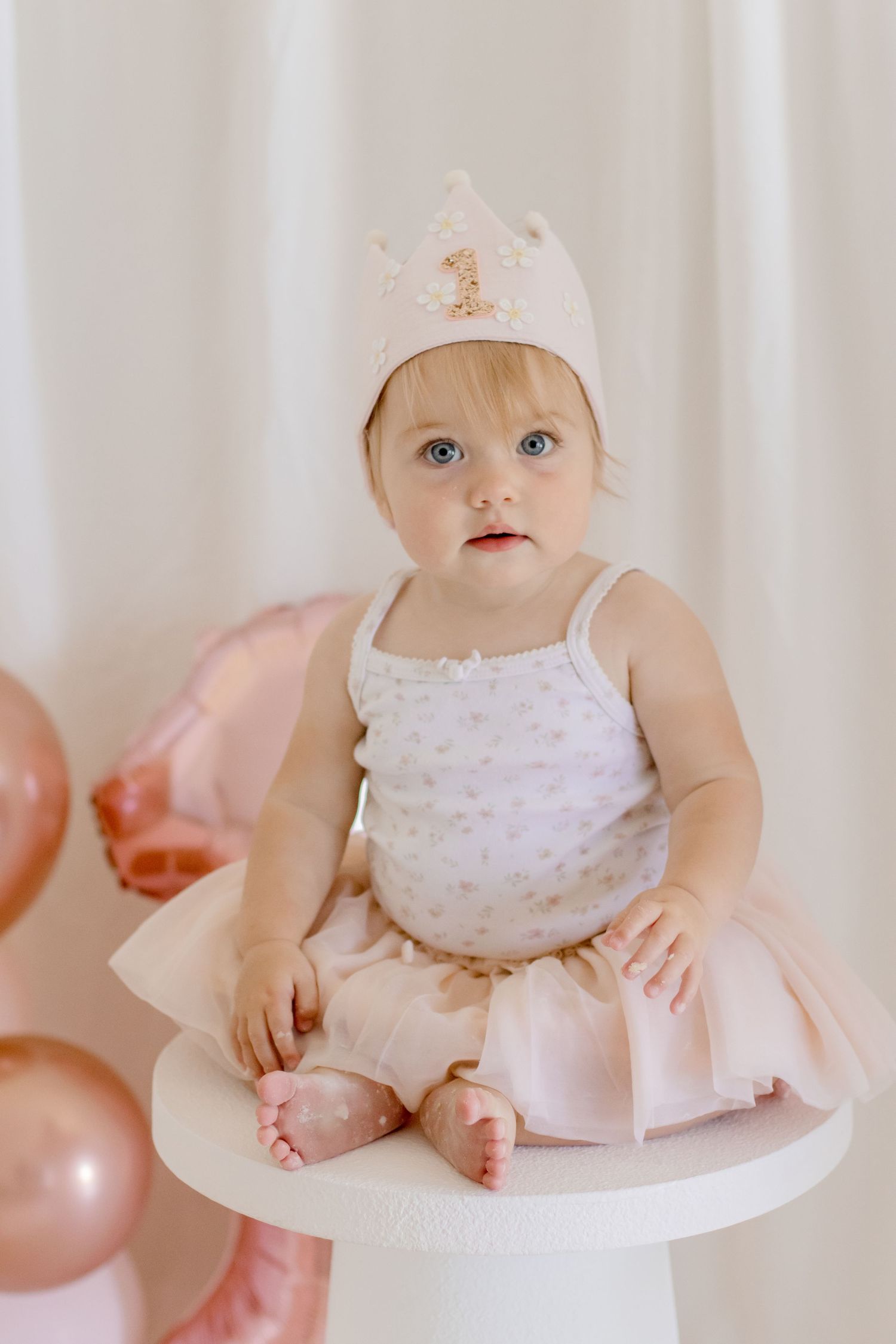 A baby in a pink tutu dress and crown sits on a white pedestal surrounded by rose gold balloons.
