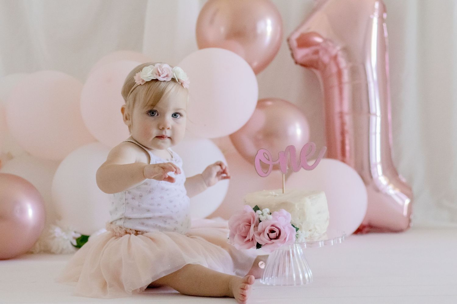 Baby reaching towards birthday cake surrounded by pink and white party decorations.