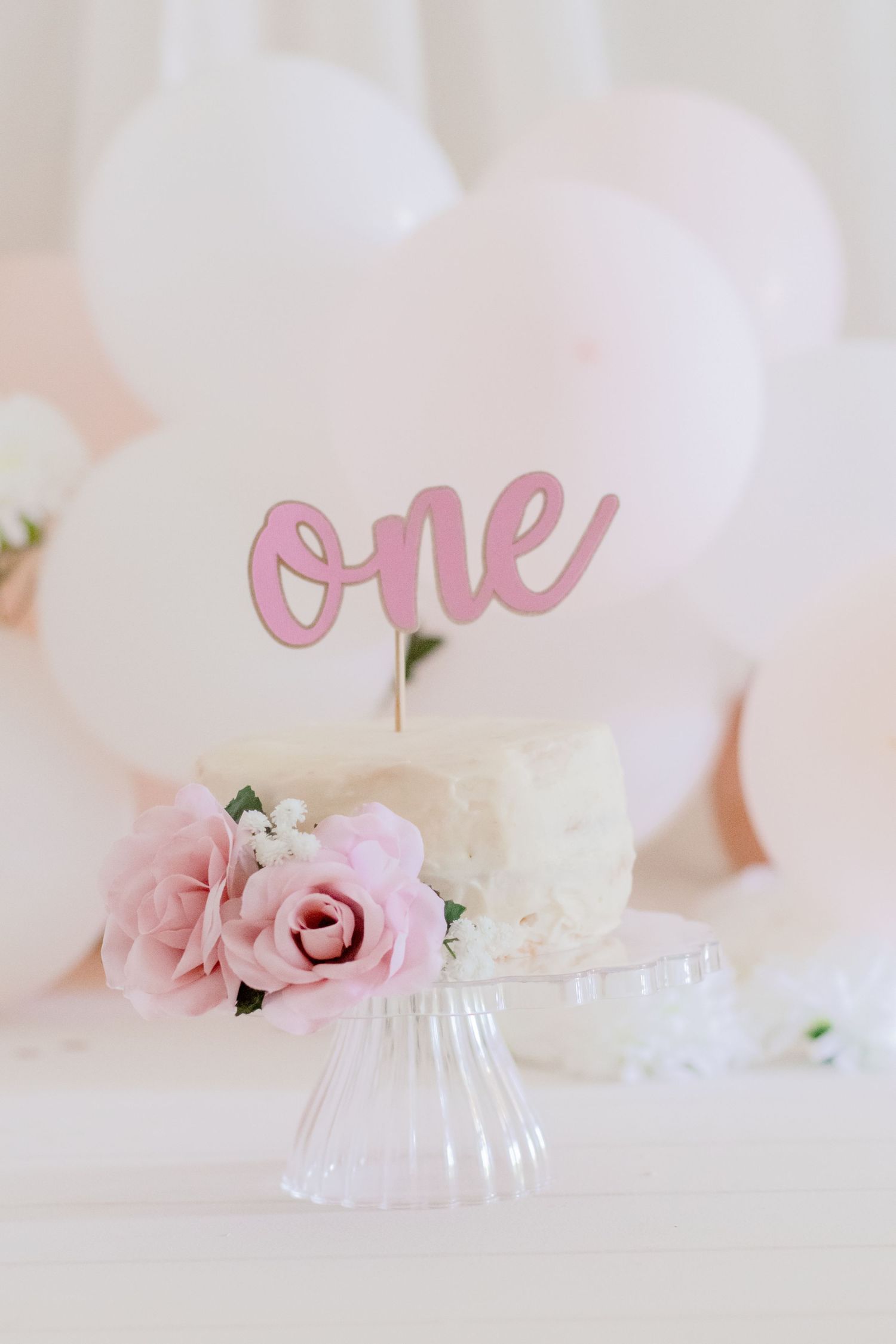 Close up of birthday cake with pink 'one' topper and roses against white balloon backdrop.