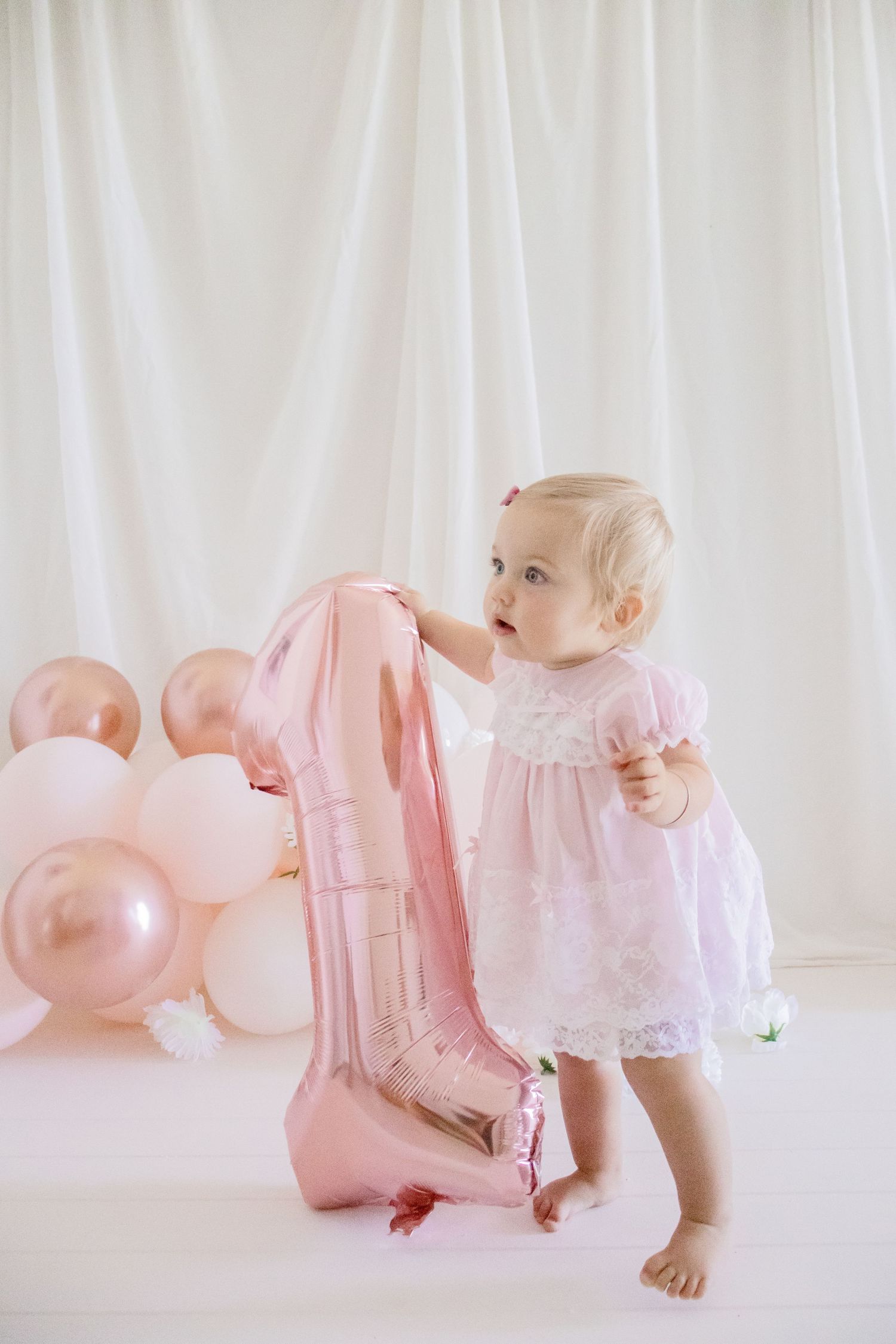 Baby in lace dress standing next to large pink number one balloon against white backdrop.