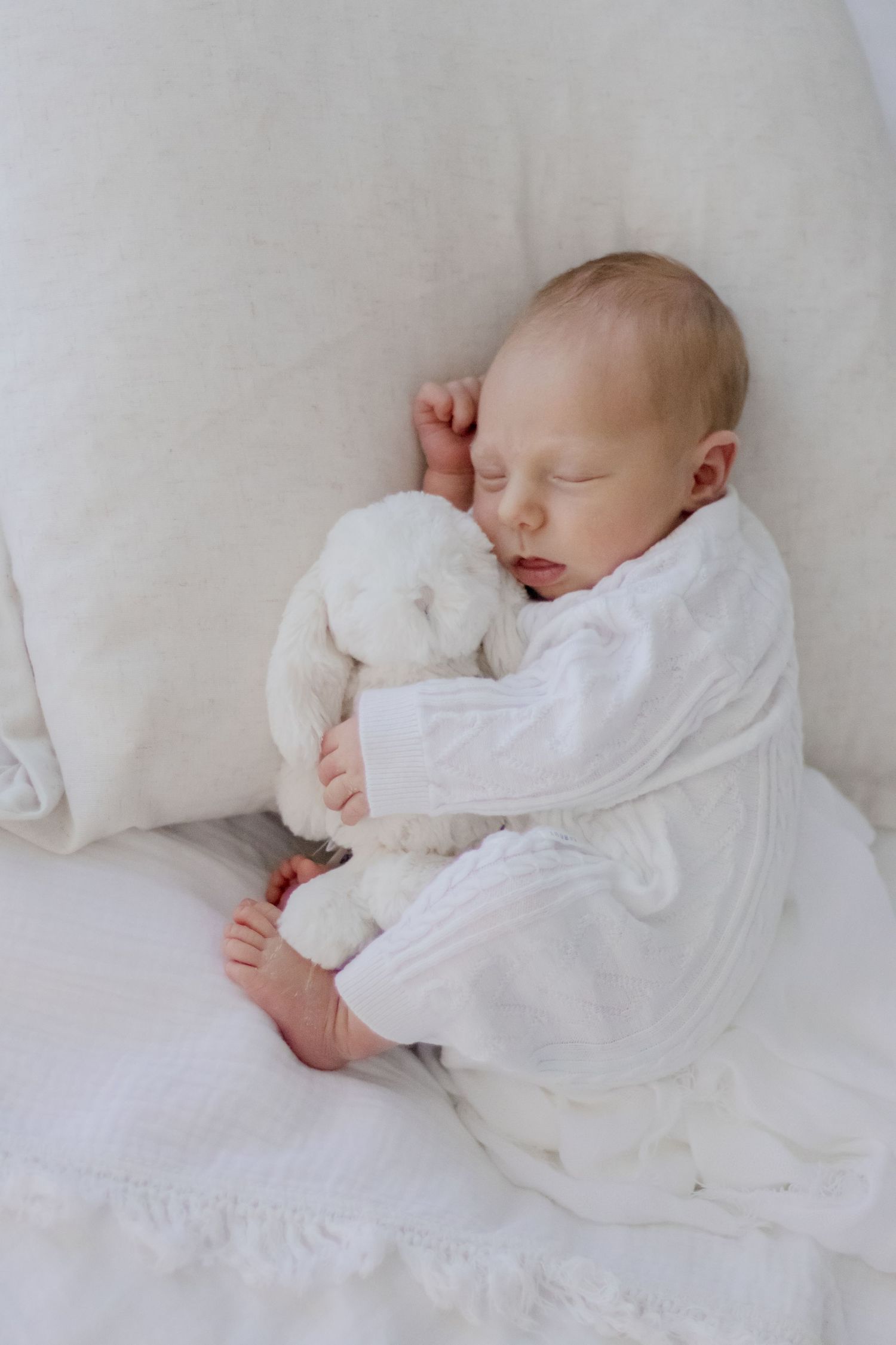 A peaceful sleeping newborn in white clothing cuddling with a plush stuffed animal.
