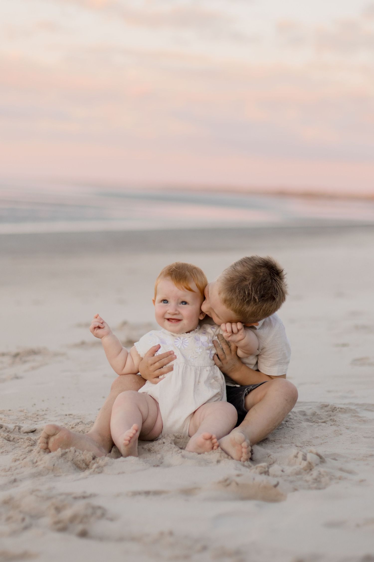Two small children sit together on a sandy beach at sunset sharing a sweet moment together.