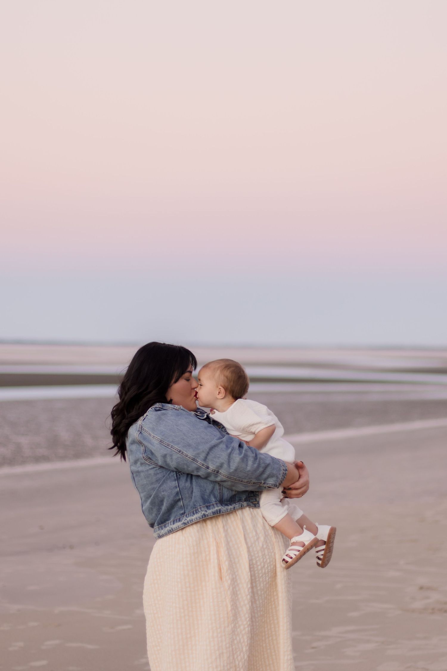 A parent in denim jacket and white skirt holds their baby on a beach at sunset with soft pink skies.