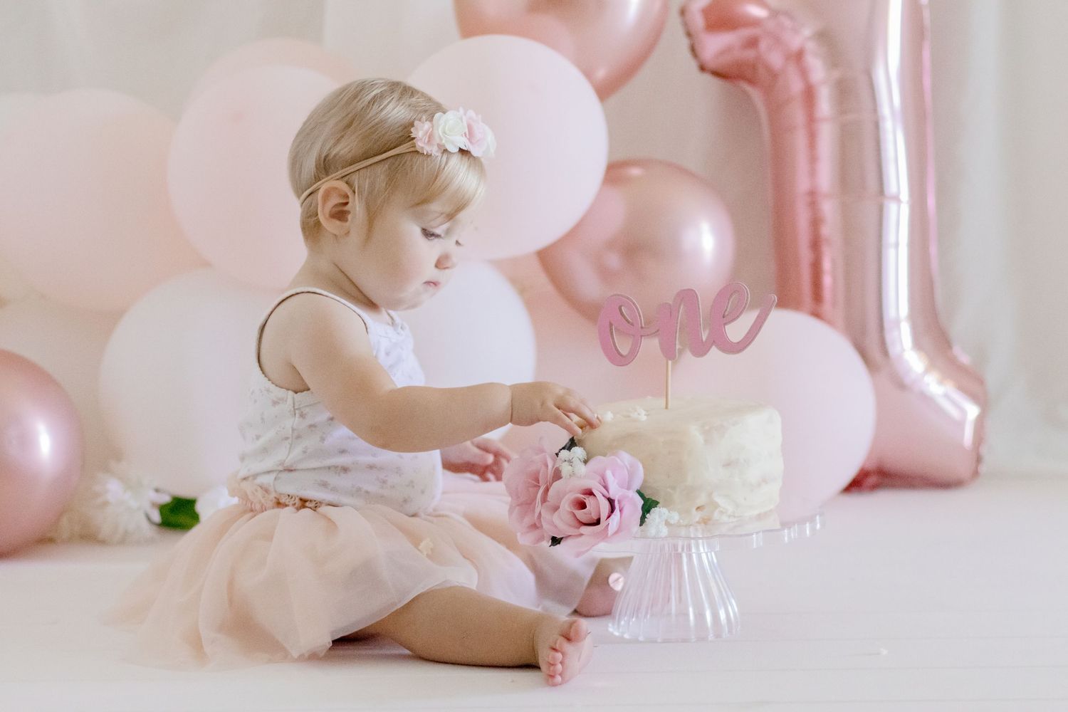 A first birthday cake smash setup with pink and white balloons, tulle dress, and floral decorations.