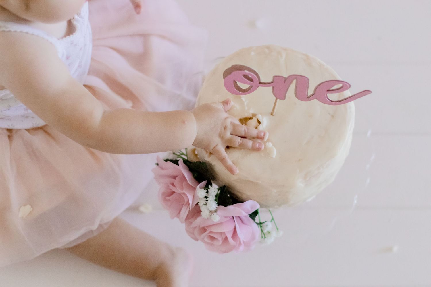 Close-up of white birthday cake with pink 'one' topper and rose decorations.