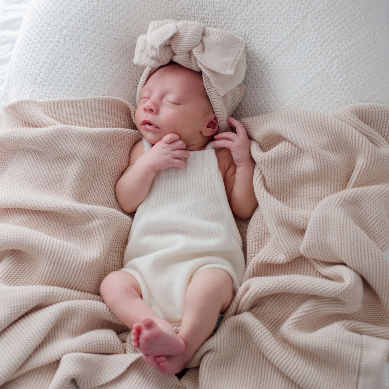 Sleeping newborn wearing cream outfit and bow headband rests on ribbed knit blanket.