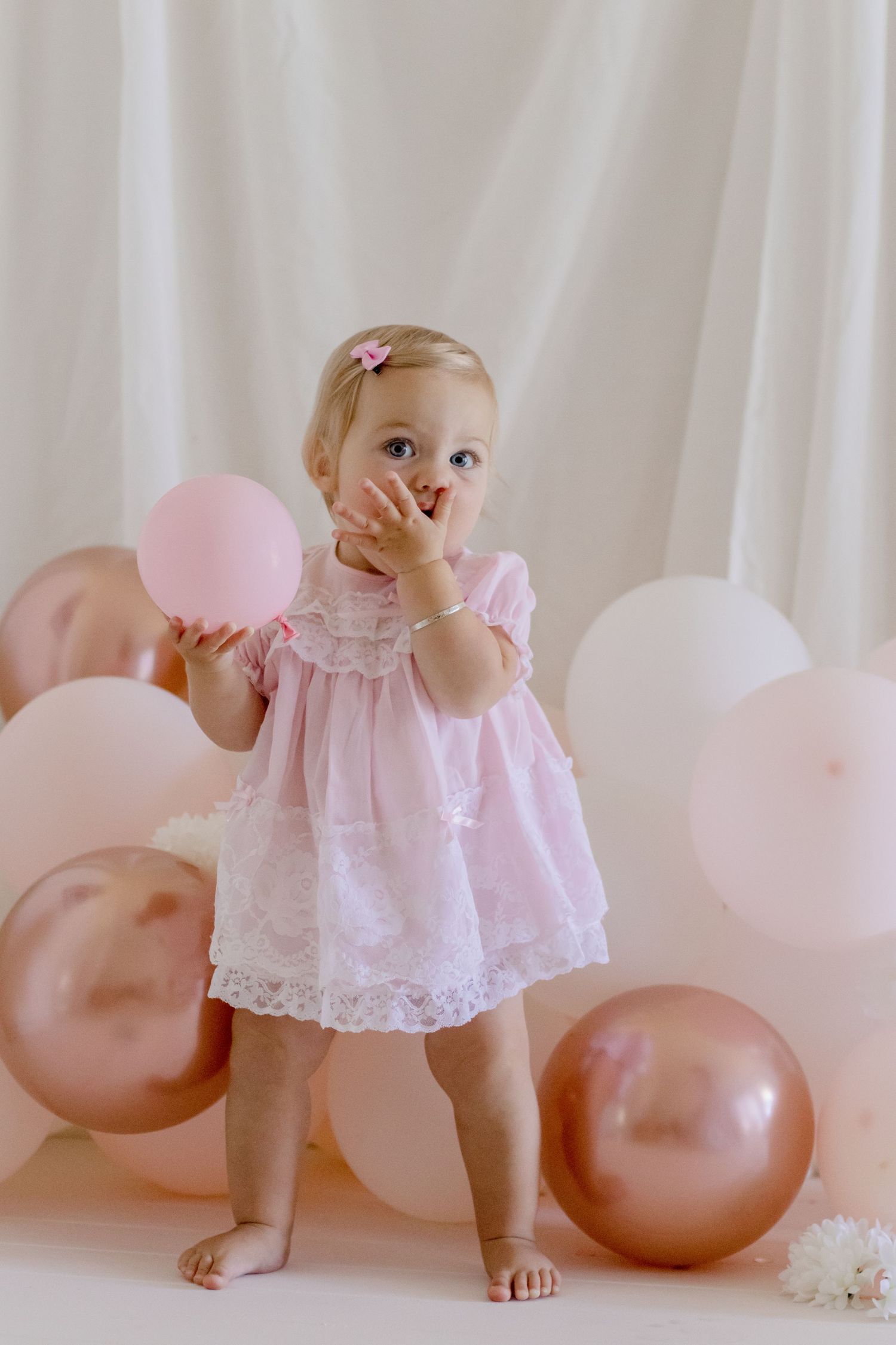 Baby in pink lace dress stands among rose gold and pink balloons.