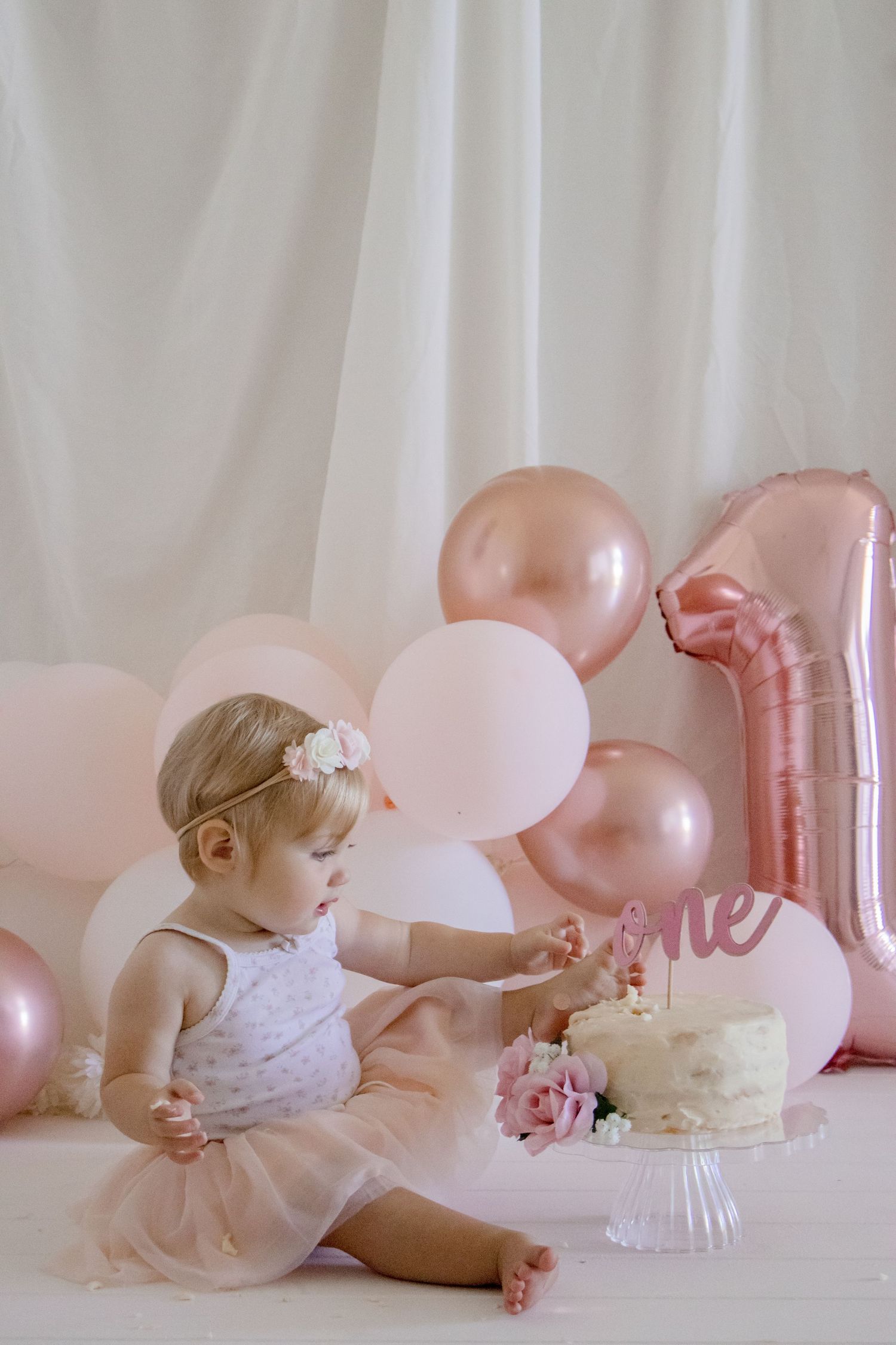 A dreamy first birthday setup with rose gold balloons, white cake, and pink roses.