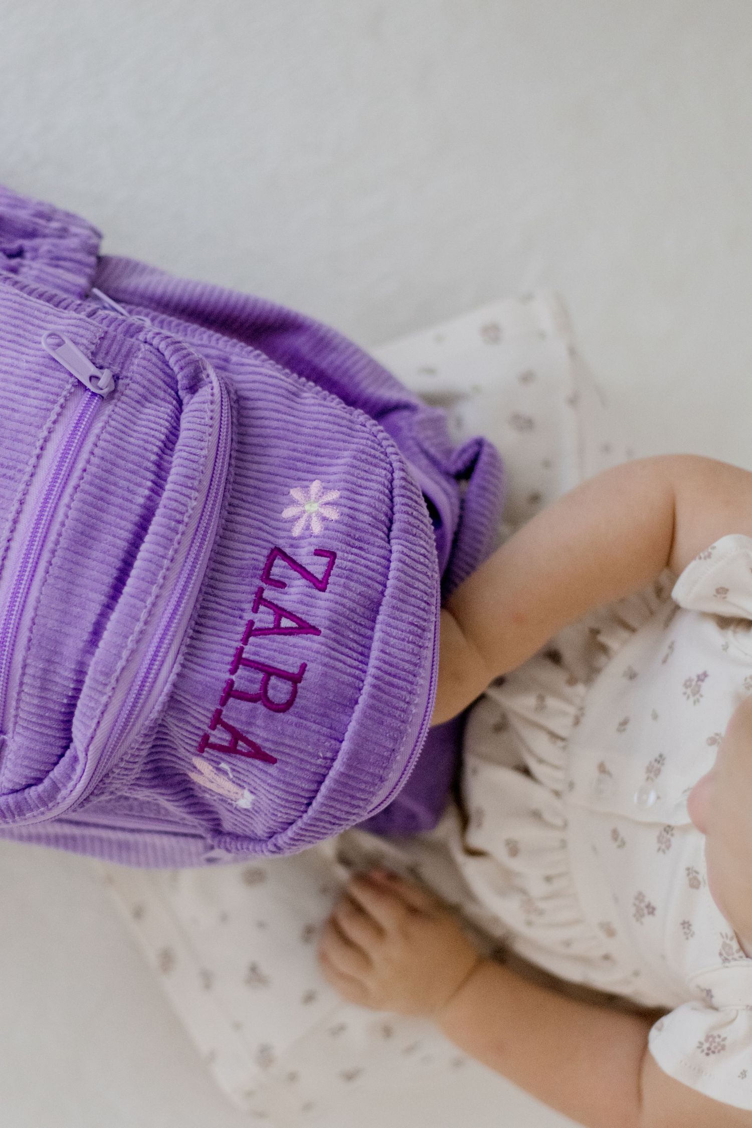 Purple corduroy backpack with floral embroidery next to white dress.