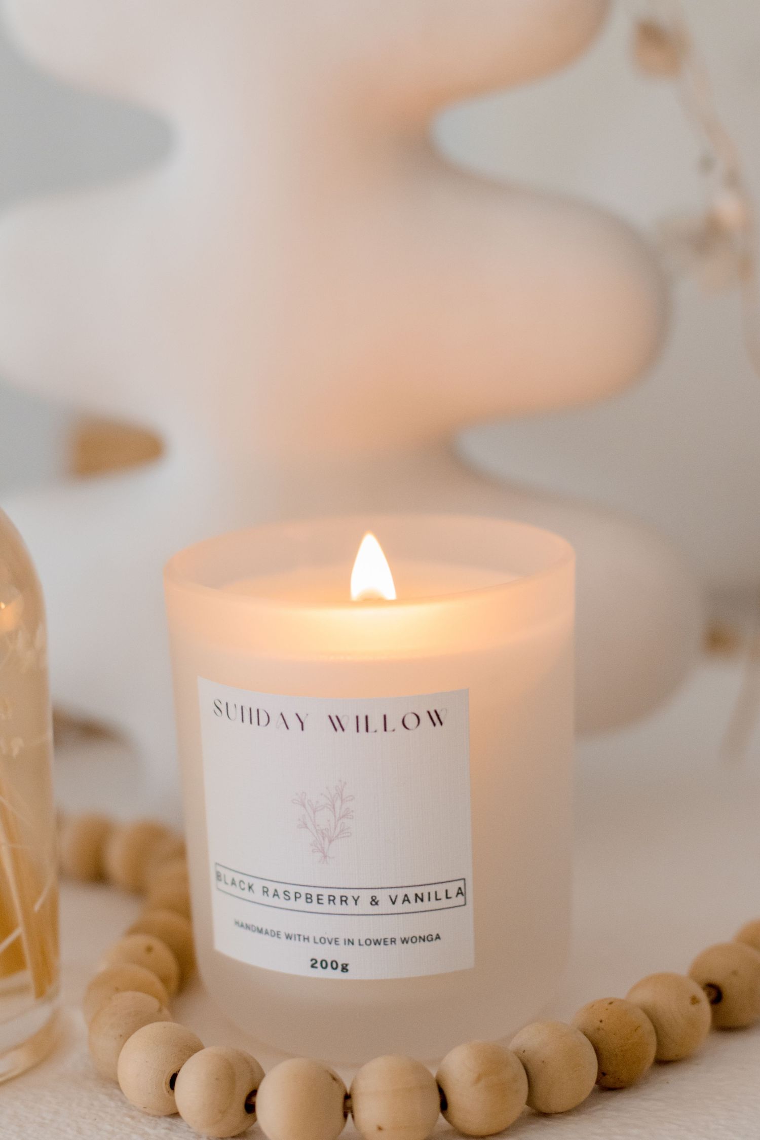 Lit Sunday Willow candle with wooden bead garland on white surface.