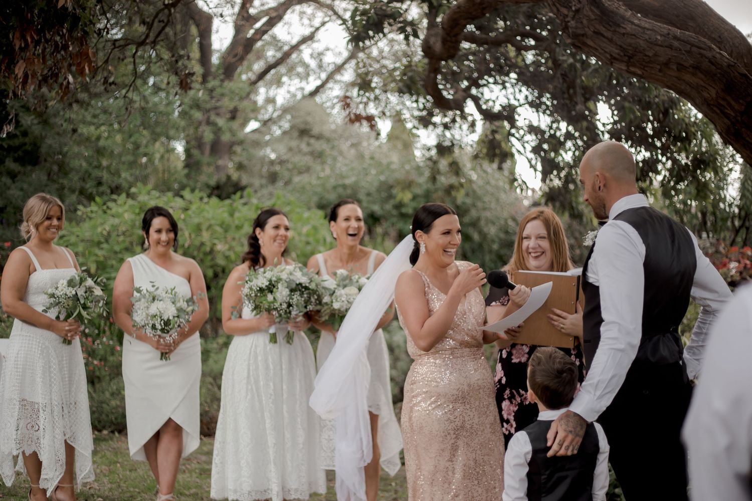 A romantic outdoor wedding ceremony takes place under tree branches with attendants in white and cream dresses.