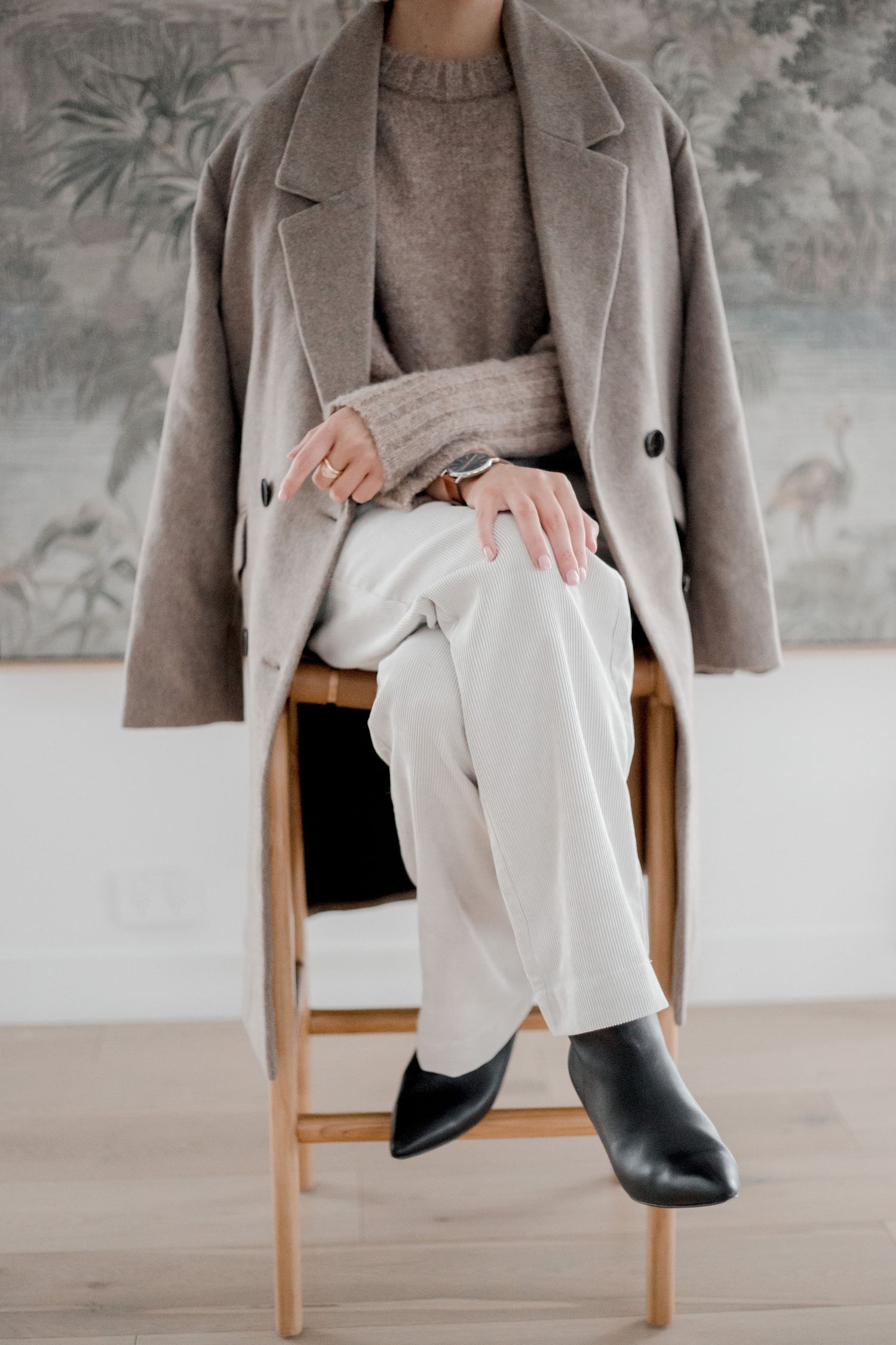 Minimalist fashion outfit featuring beige coat, white pants and black boots while seated on wooden stool.