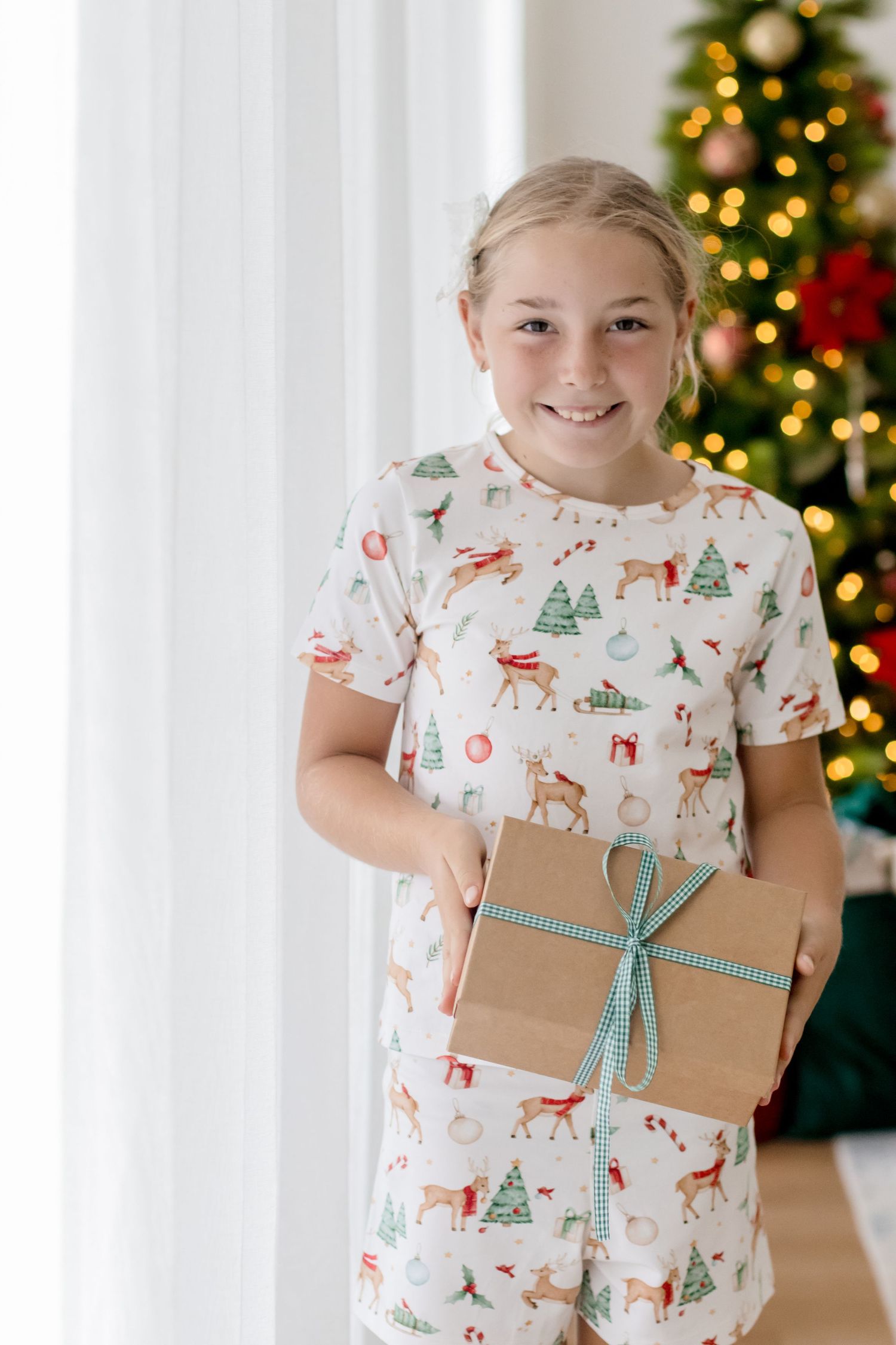 Child wearing festive holiday pajamas holds wrapped Christmas present with teal ribbon against decorated tree background.