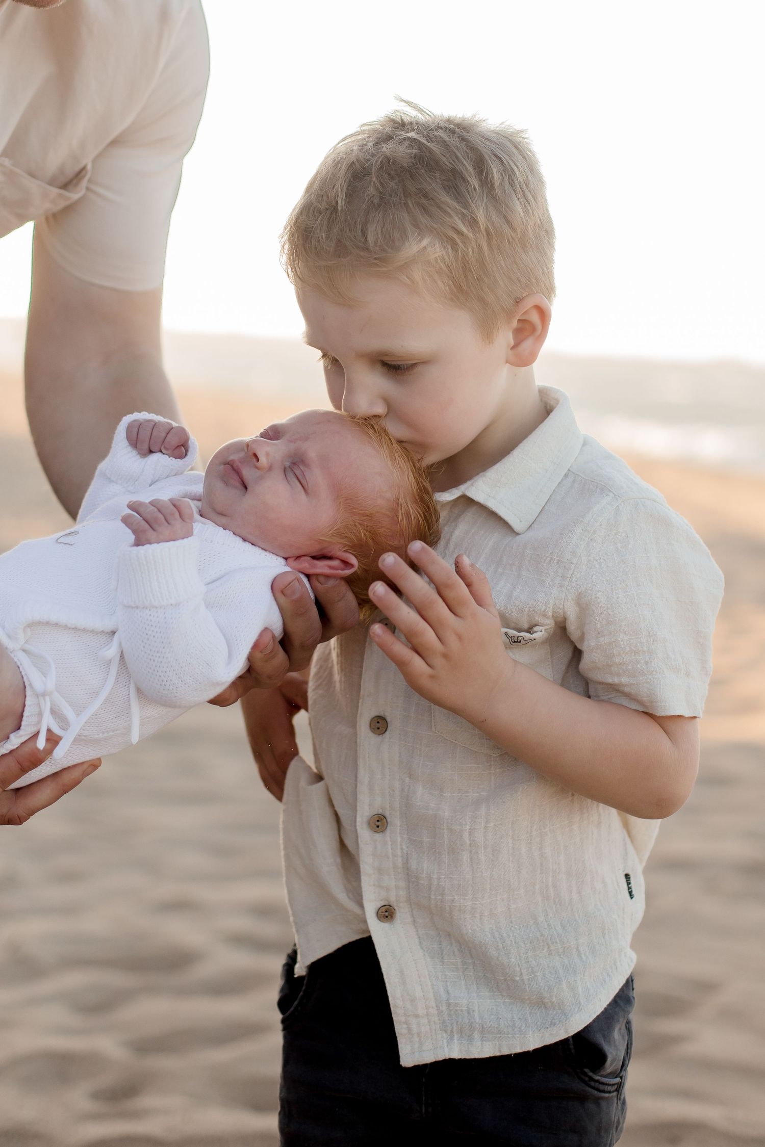 Sibling bonding moment as older child gently kisses newborn in white outfit on beach at sunset.