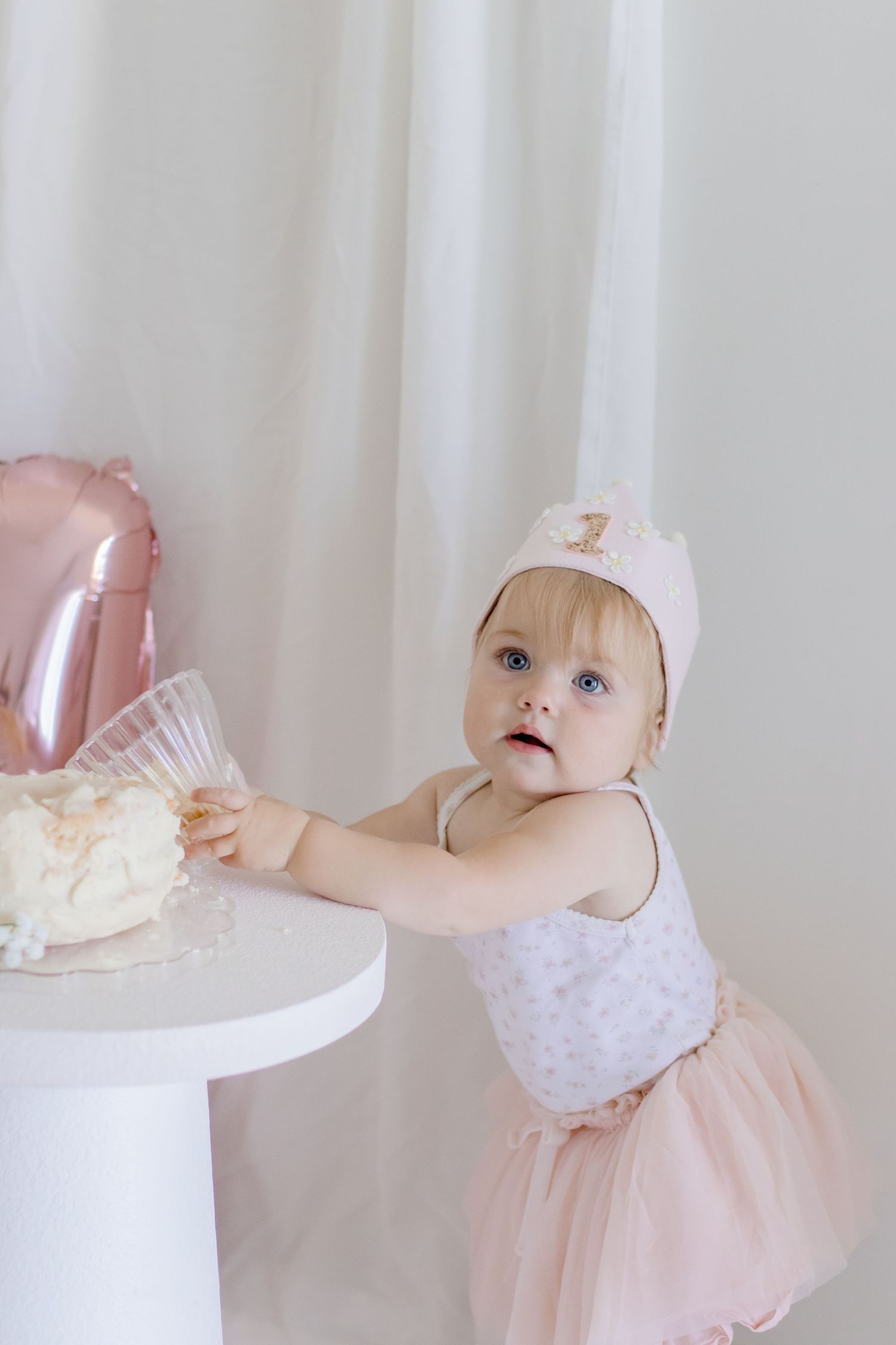 Baby in pink tulle dress and crown reaching for cake at first birthday celebration with white and pink decor.