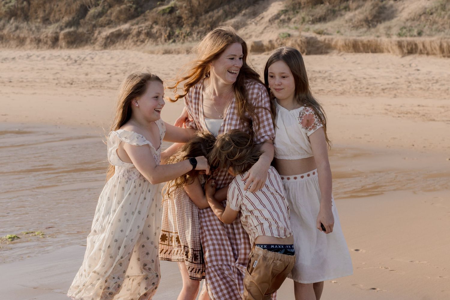 Group in vintage-style dresses enjoying a playful moment together on a beach with dunes in the background.