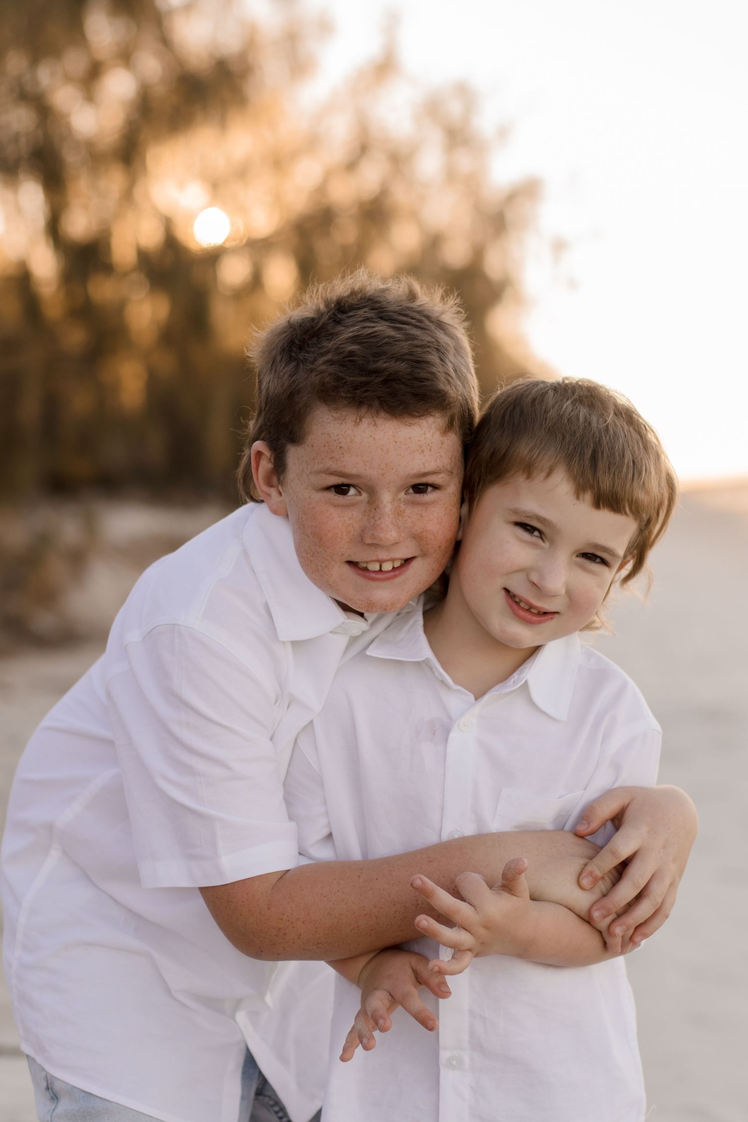 Two children in white shirts embrace and smile while posing outdoors at sunset.