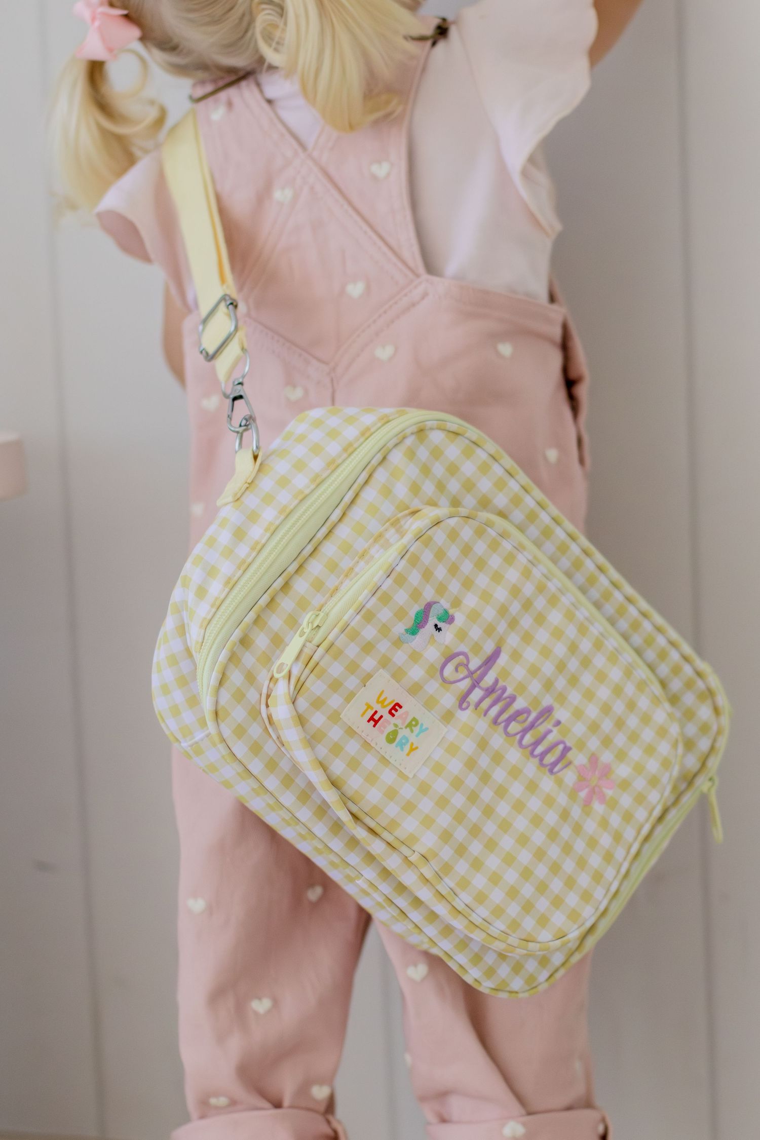 Toddler girl wearing pink heart overalls with a personalized yellow gingham mini backpack named Amelia.