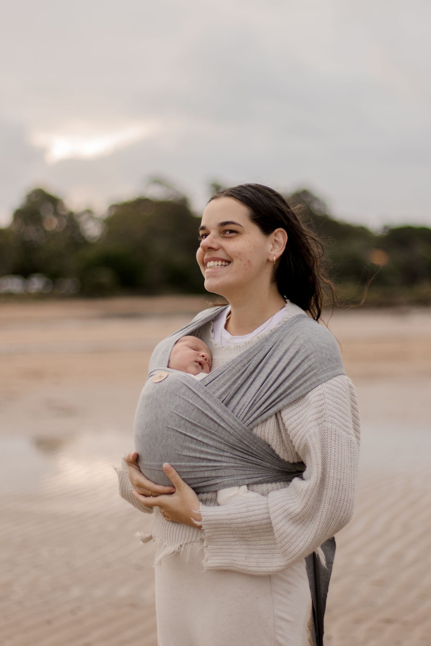Parent wearing cream sweater carries infant in grey baby wrap carrier at beach during sunset.
