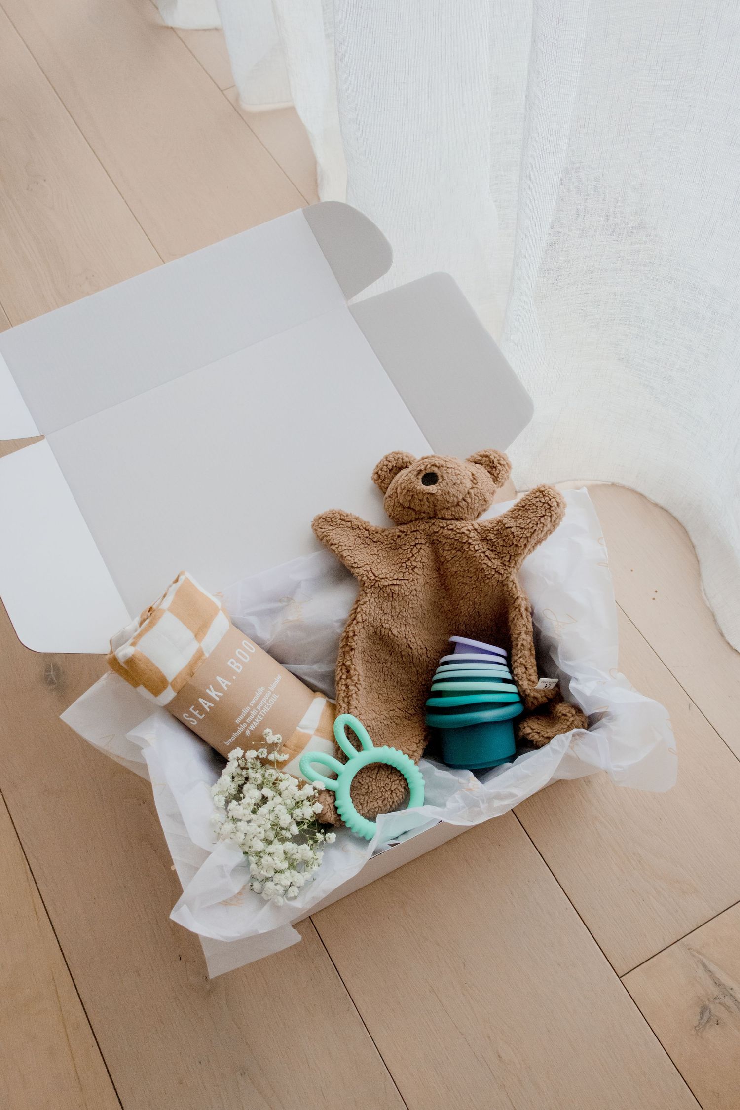 A stylish gift box containing a teddy bear, socks, baby items and white flowers on wooden flooring.
