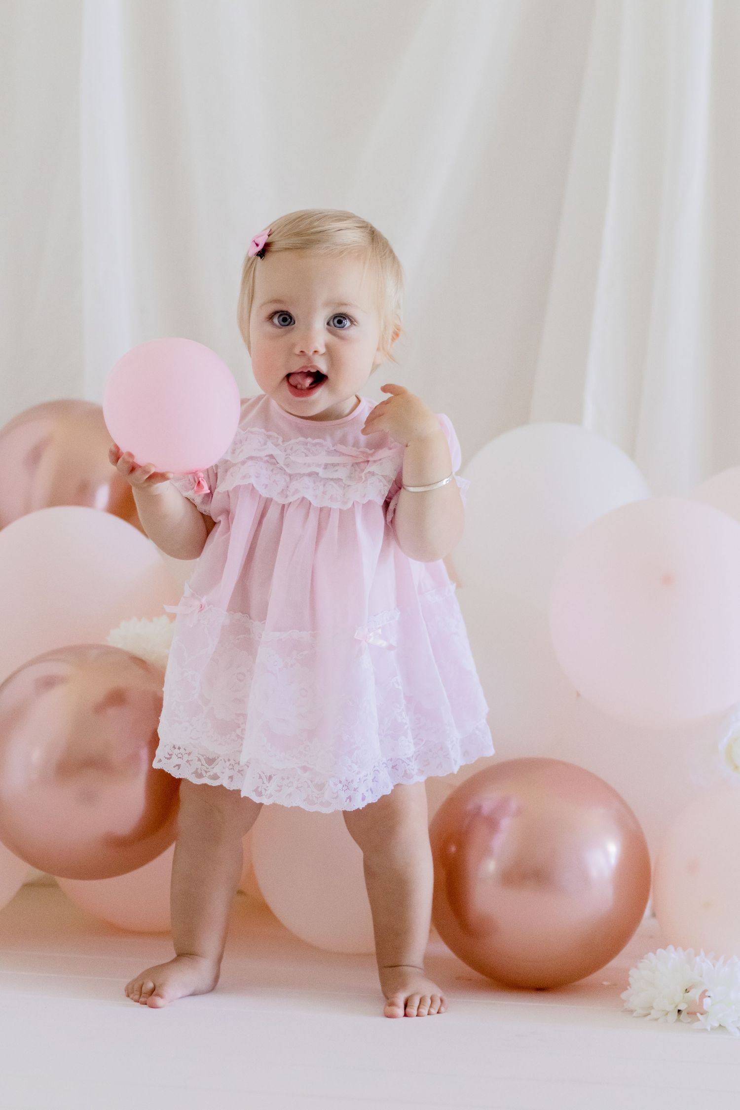A toddler in a pink lace dress stands barefoot surrounded by pink and rose gold balloons against a white backdrop.