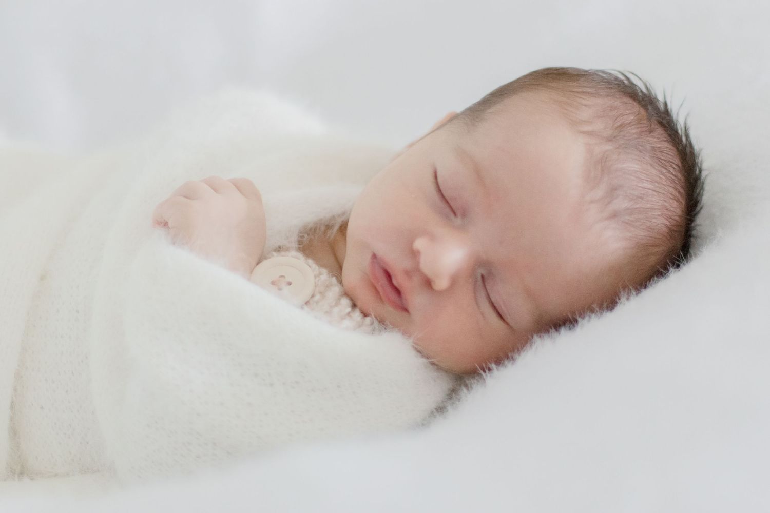 Peaceful sleeping newborn wrapped in white blanket against light background.