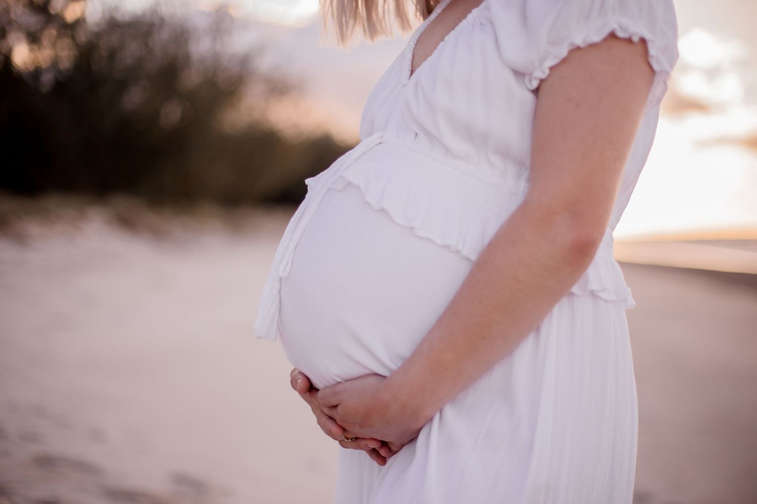 Side profile of pregnant woman in white dress holding baby bump on beach at sunset.