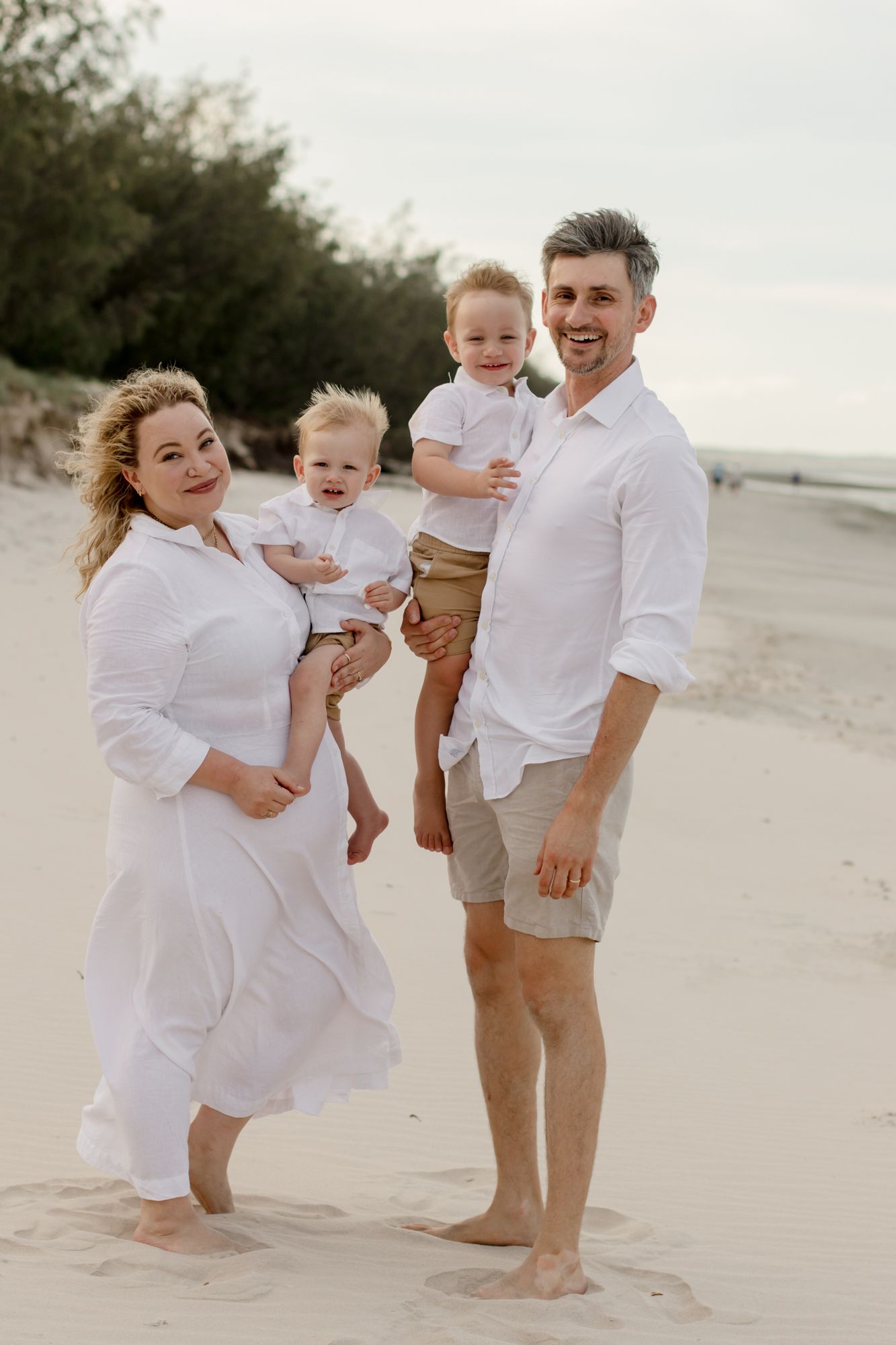 A family dressed in white and beige poses together on a sandy beach.