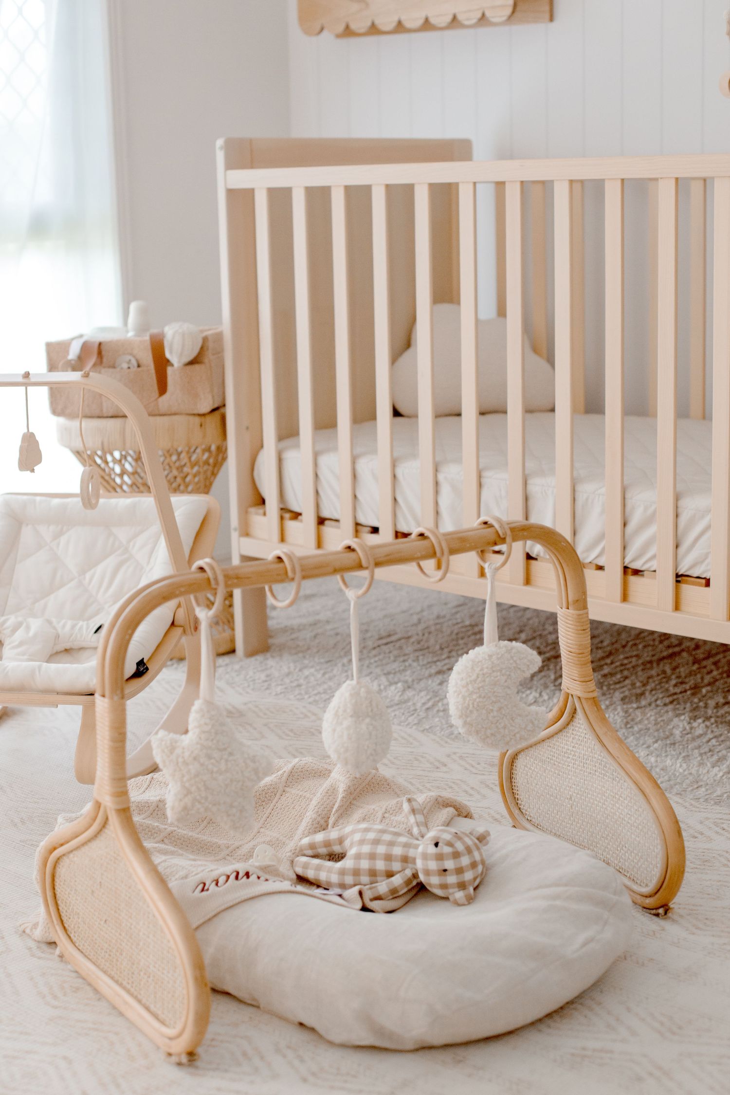 Wooden baby crib with white bedding and natural wooden play gym with hanging soft toys and cushions.