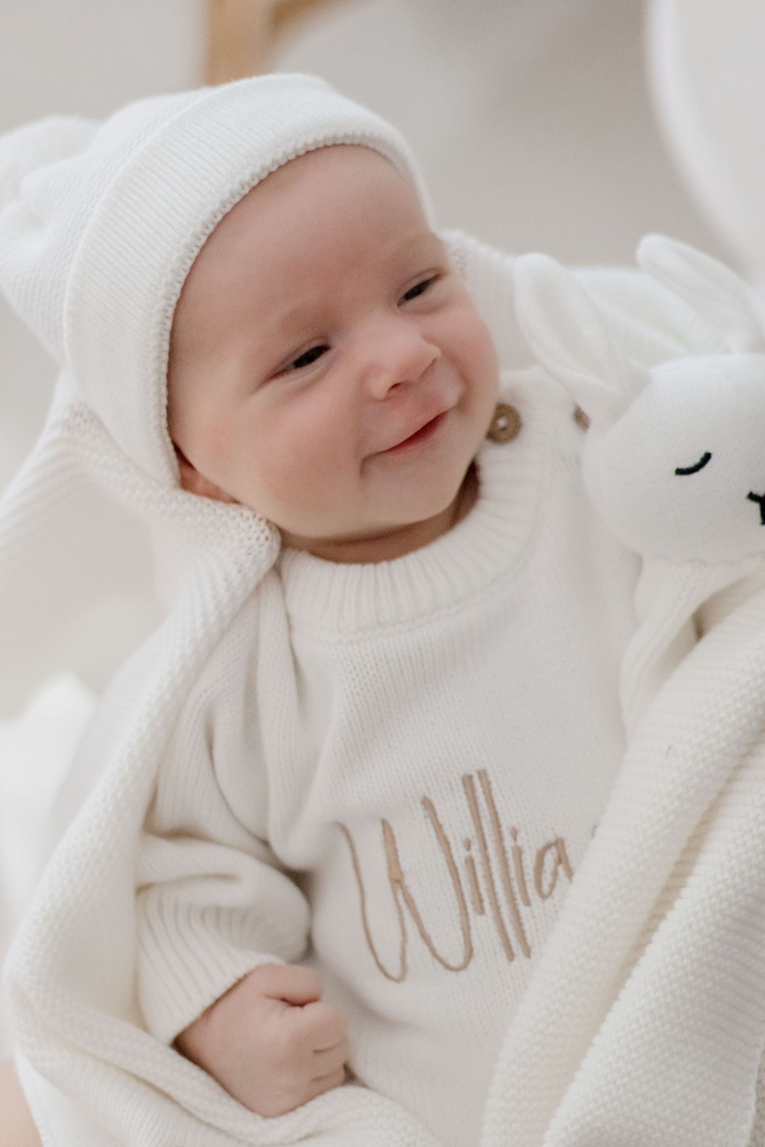 Baby in white winter outfit smiling while lying next to plush toy on white bedding.