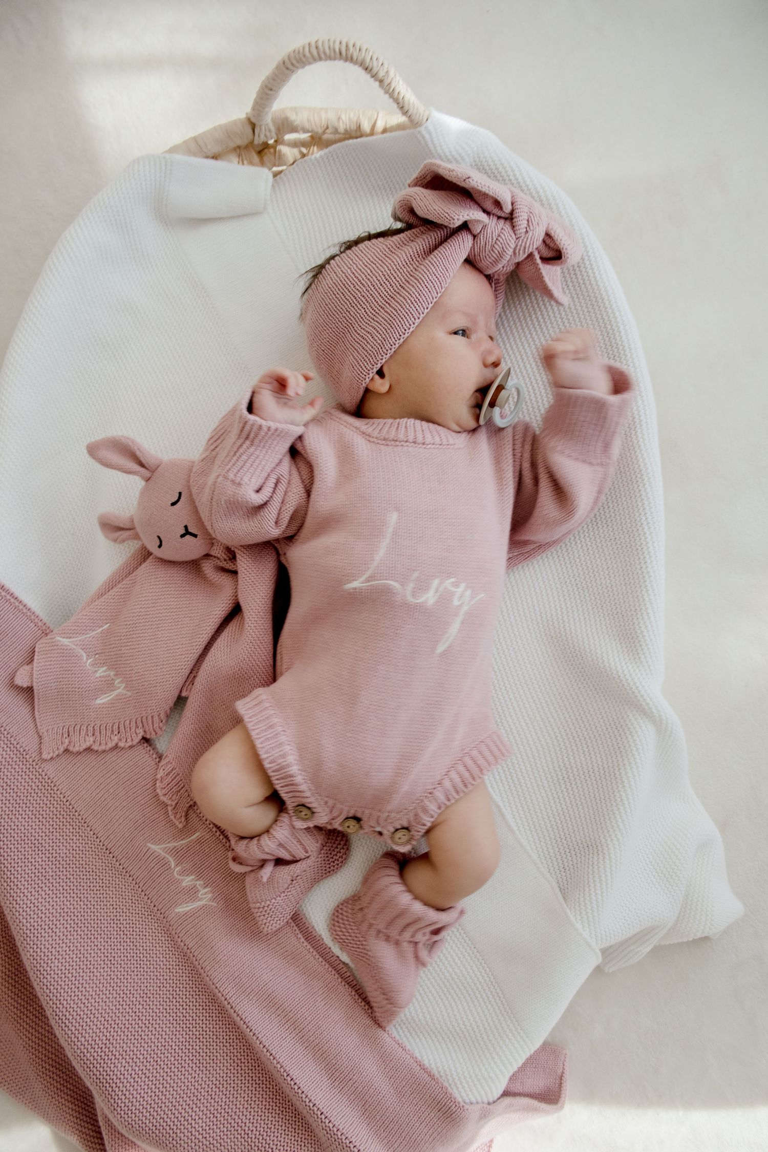 Baby lying in moses basket wearing pink knitted outfit and headband with matching stuffed bunny toy.