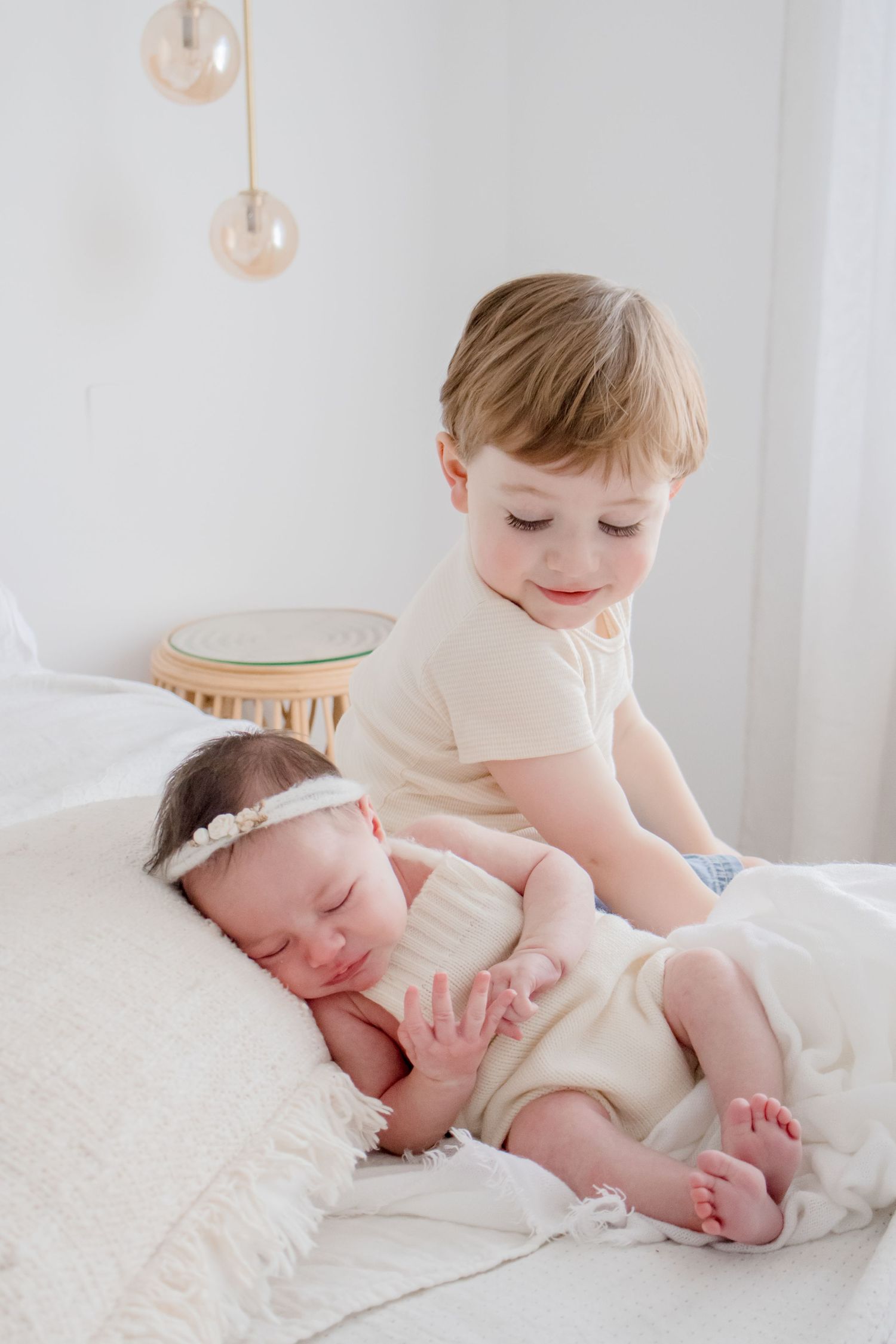 Siblings share a tender moment as they rest together on white bedding with pendant lights above.