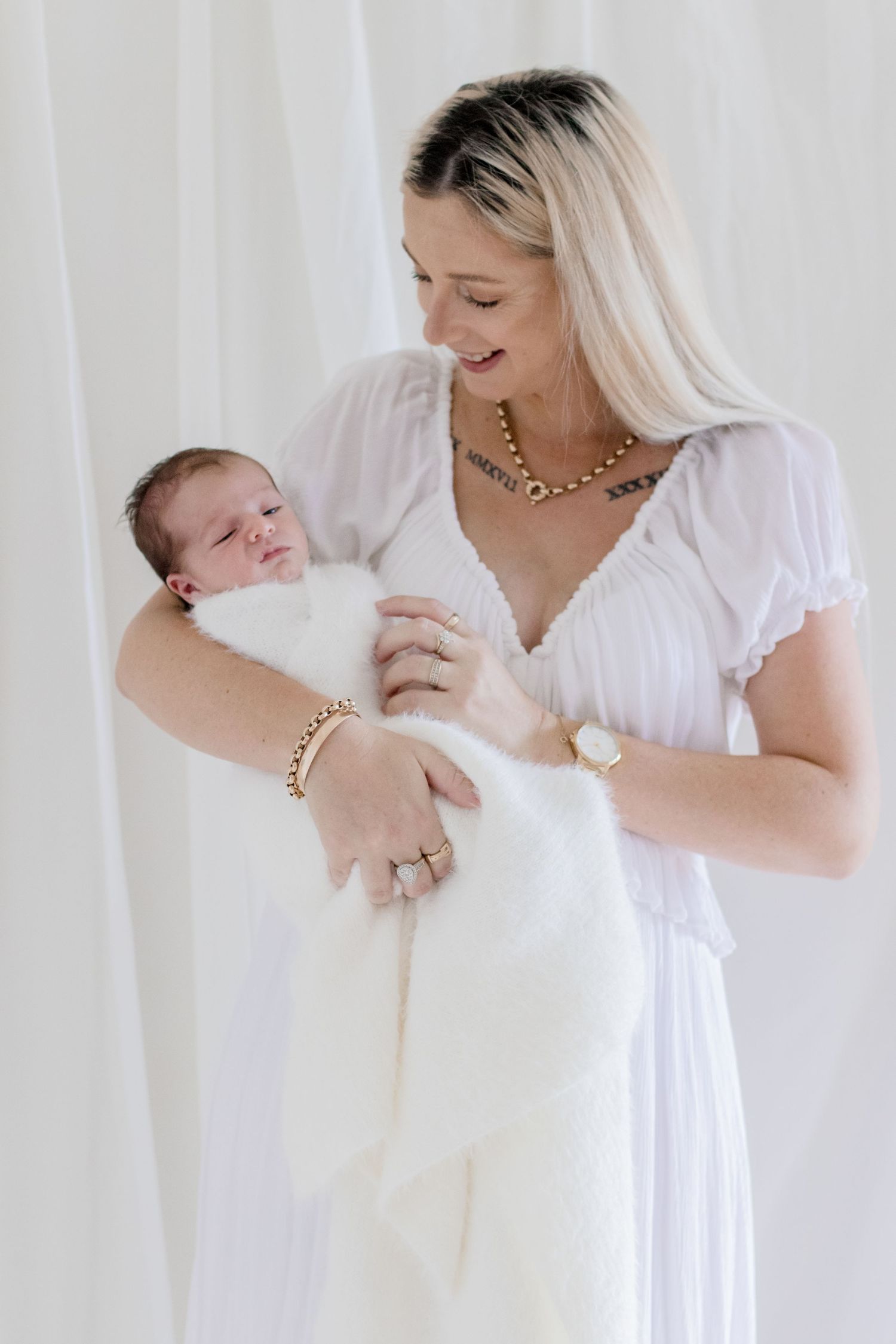 Mother in white dress holding newborn baby wrapped in soft white blanket against light background.