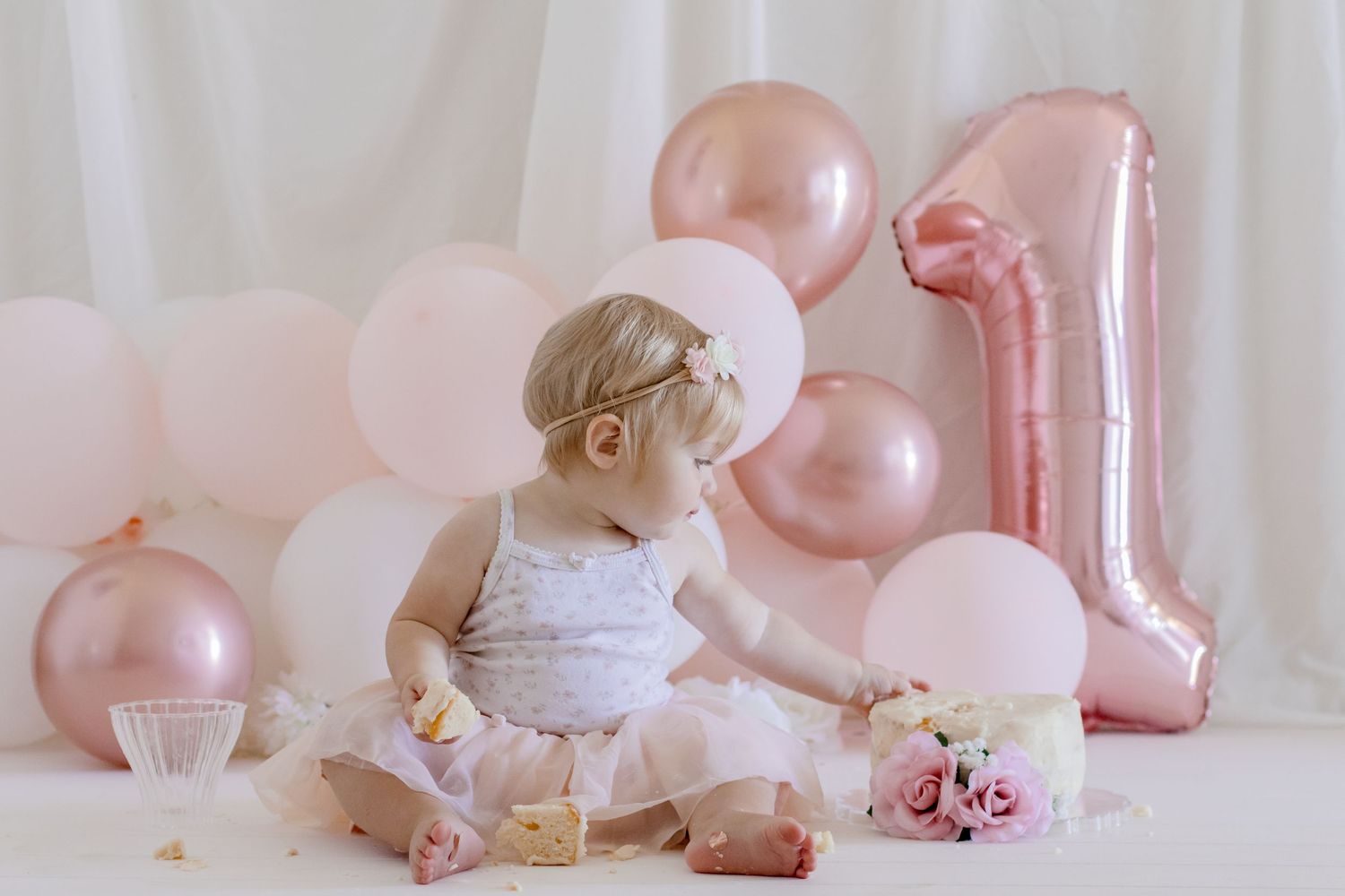 Baby in party dress sitting with pink balloons and roses for first birthday.
