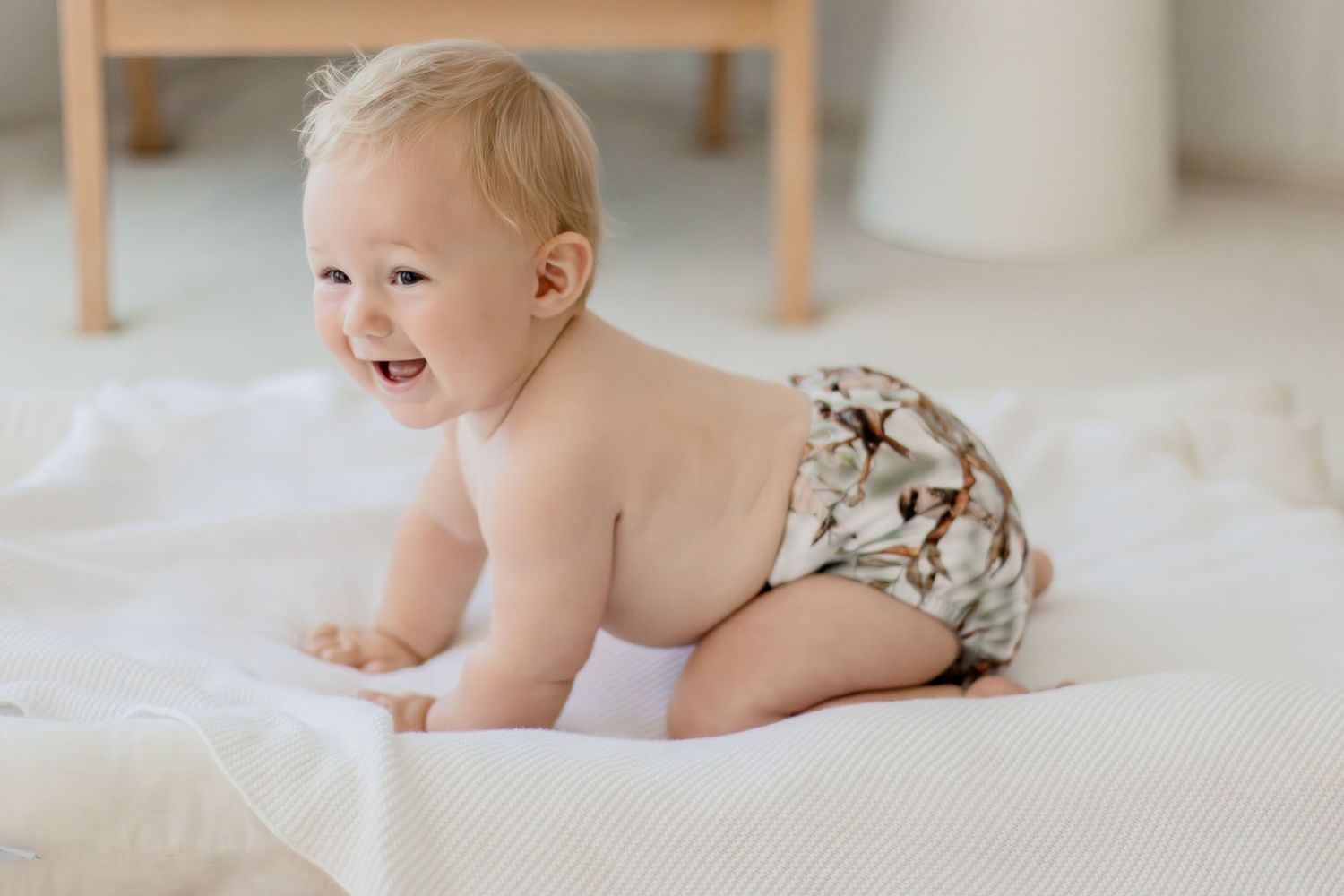 Smiling baby crawling on white blanket wearing a floral cloth diaper in a bright, airy room.