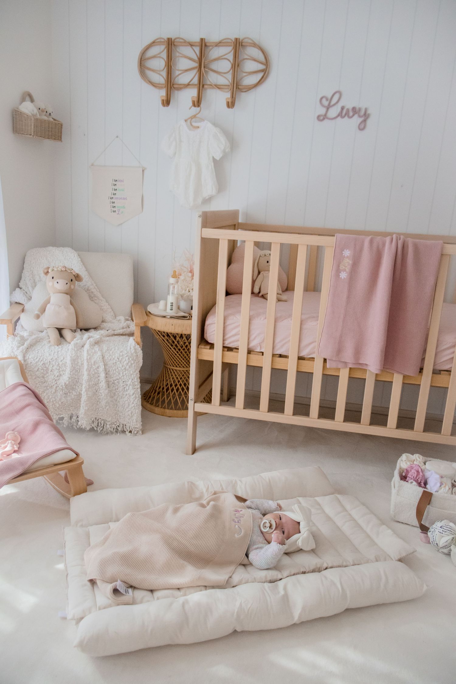 A cozy nursery corner with wooden crib, pink blanket, rattan decor, and soft white bedding.