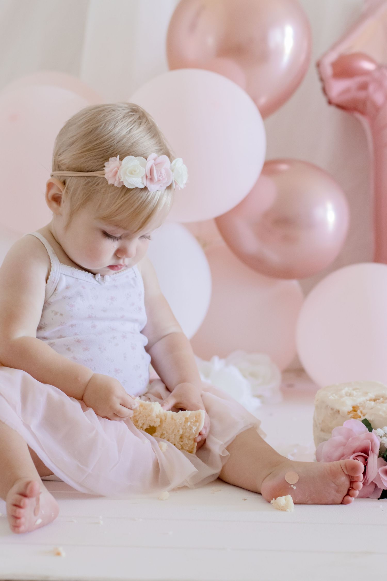 Baby wearing floral crown and white dress holding food against pink balloon backdrop.
