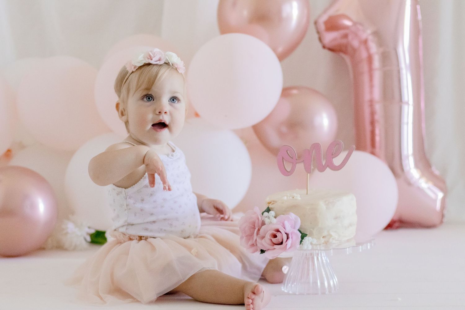 Baby in tulle dress exploring birthday cake with pink balloon backdrop and number one decoration.