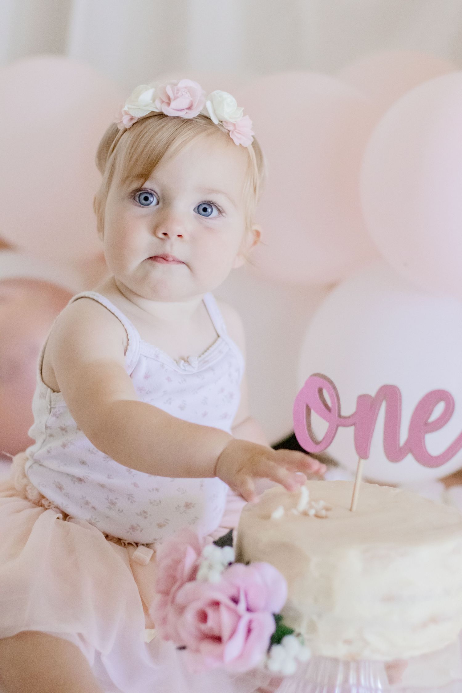 Baby in white dress with floral crown poses next to first birthday cake.