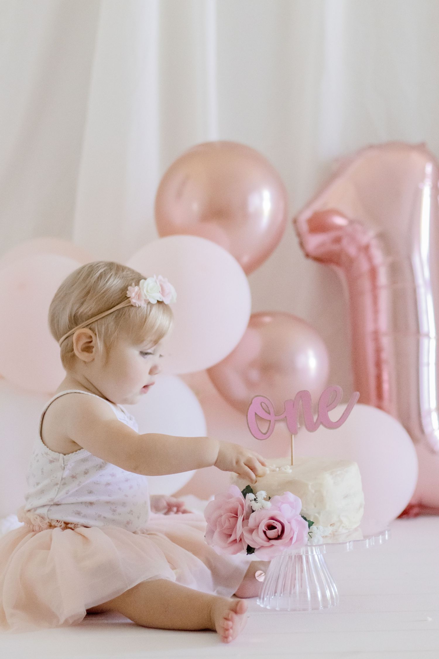 Baby in white dress examining cake during first birthday photoshoot with pink balloons.