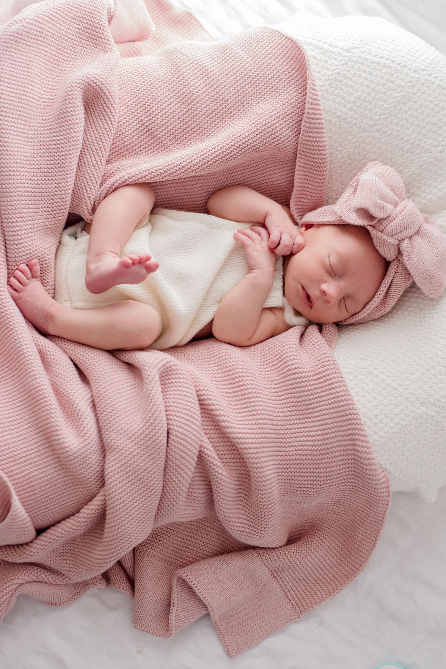 Peaceful newborn sleeps in cream outfit on pink knit blanket with matching bow headband.