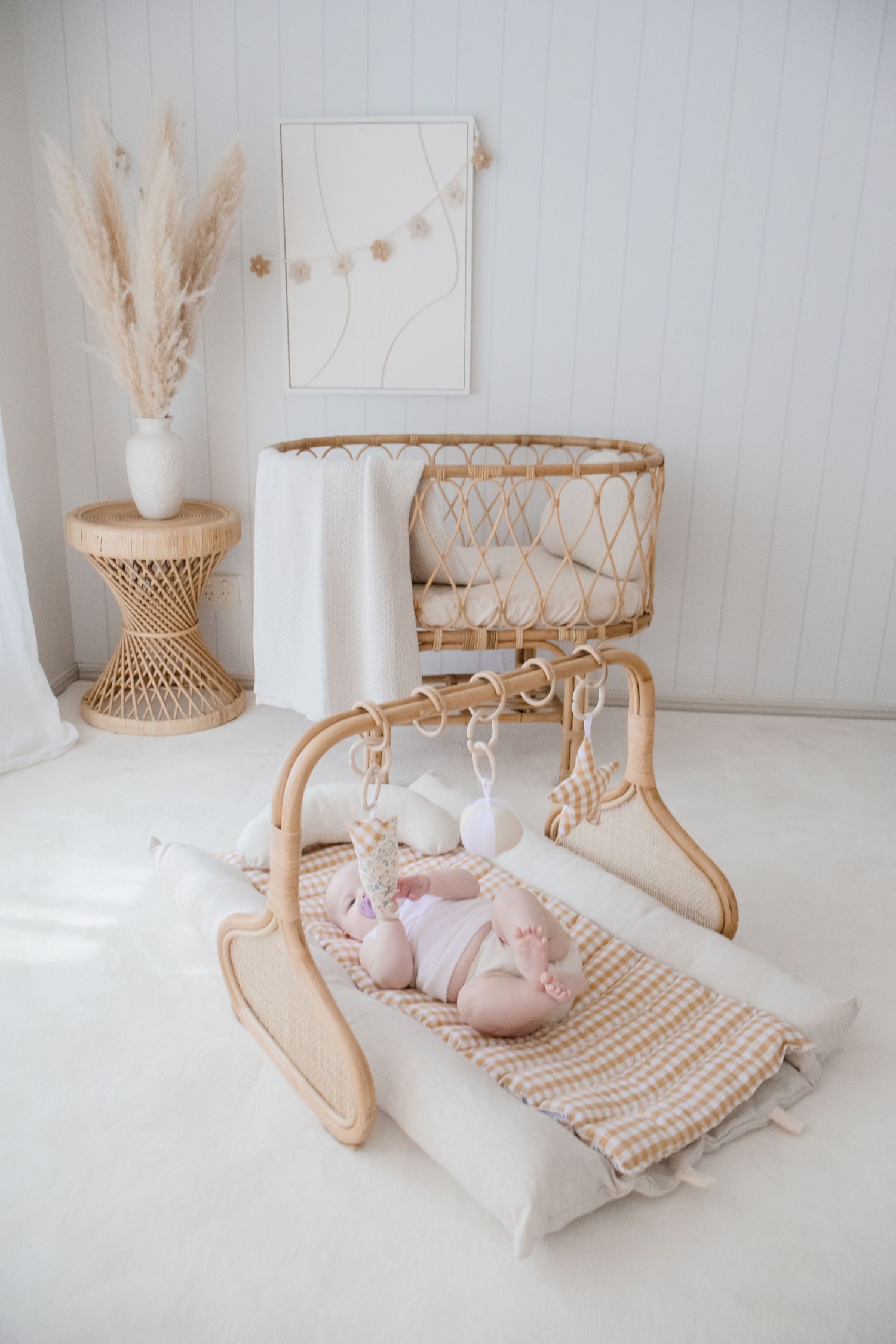Neutral nursery decor with wooden baby gym, pampas grass, and pink baby shoes on a cream striped mat.