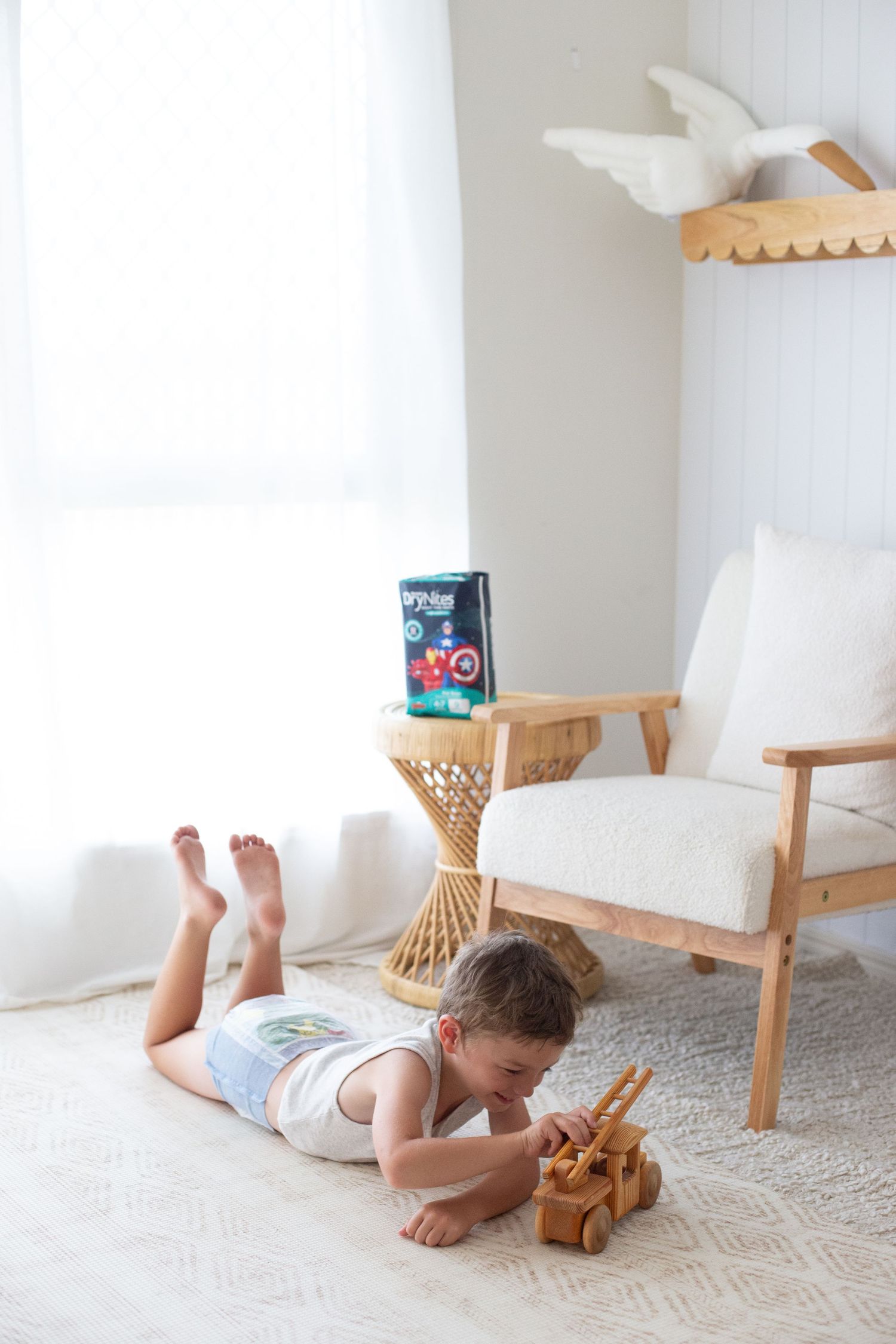 Child playing with wooden toy truck while lying on carpet near white armchair.