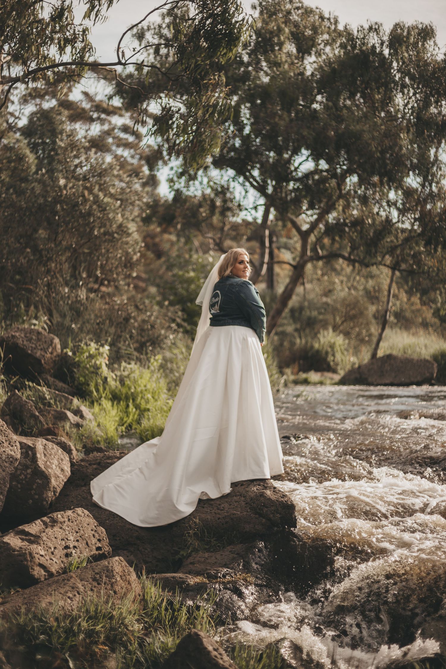 A flowing white skirt contrasts against dark rocks and rushing water in a natural outdoor setting beneath eucalyptus trees.