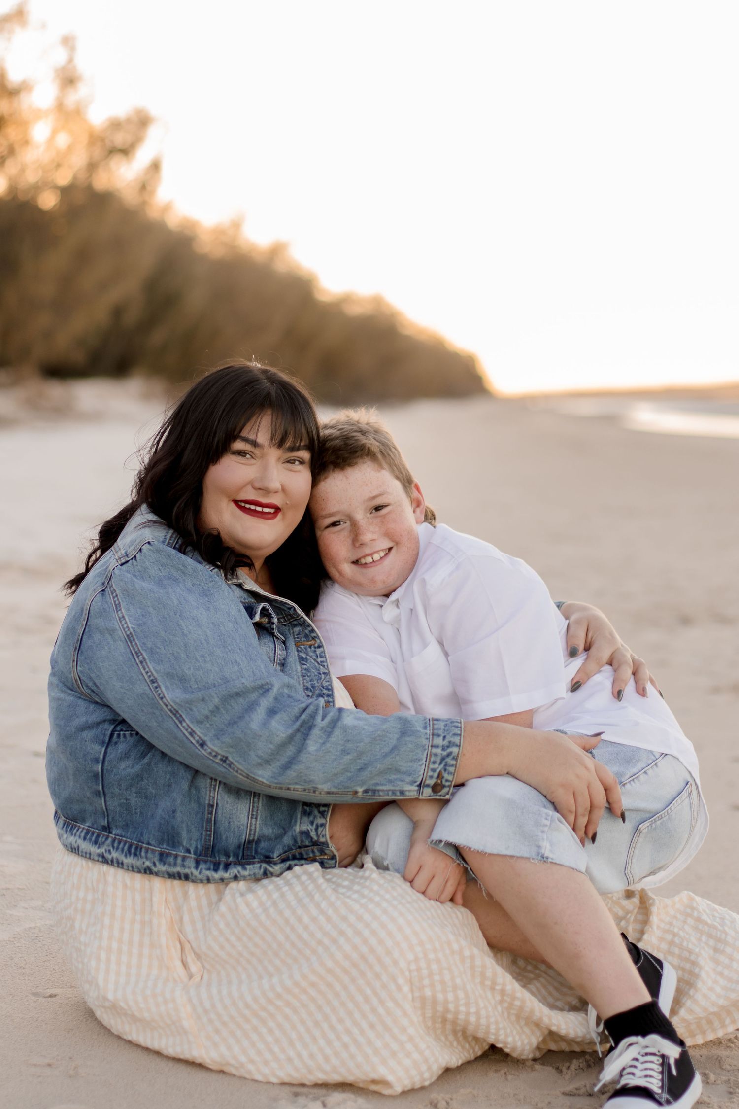 A heartwarming beach portrait shows two people embracing while sitting on sand at sunset.