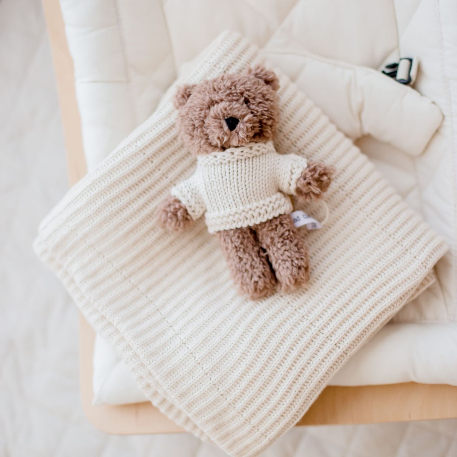 A brown teddy bear wearing a white knit sweater sits on a cozy cream-colored knit blanket.